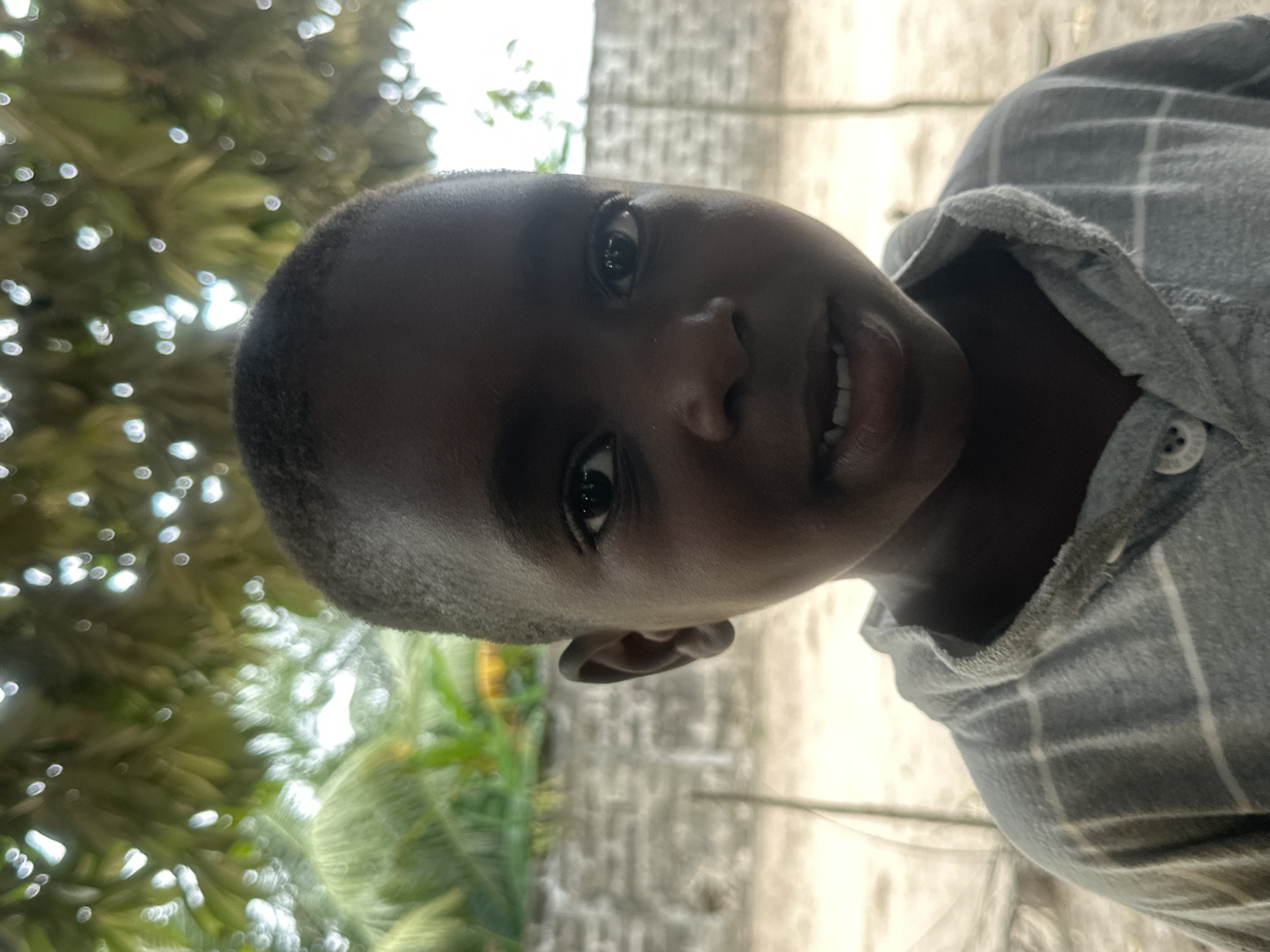 A close-up, head-and-shoulders portrait shows a young Black child facing the camera. The child has dark skin, short dark hair, and dark eyes. They are wearing a grey collared shirt with a subtle plaid pattern. The background is out of focus, with greenery and what appears to be a brick wall visible. Sunlight dapples through leaves overhead. The setting is outdoors, likely in a rural or semi-urban area of Badagry, Nigeria, suggested by the tropical foliage and simple construction. No specific actions or interactions are depicted; the image is a static portrait.