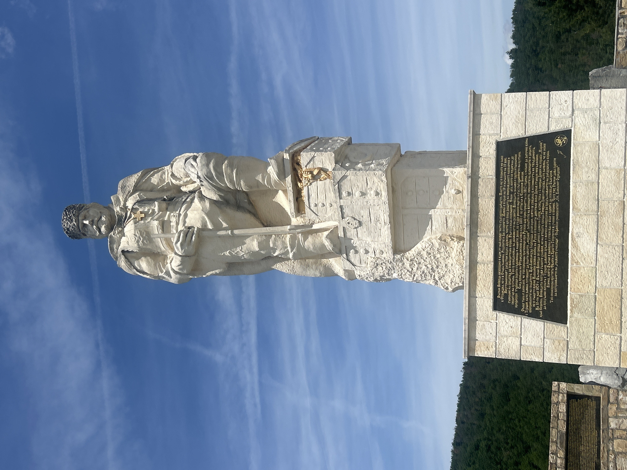 The media file displays a large stone monument of a male figure, standing against a clear blue sky with some wispy clouds and a visible contrail. The statue, made of light-colored stone, depicts a determined-looking man dressed in historical attire, including a textured cap, a buttoned jacket, and trousers. A cross pendant hangs around his neck, and he holds a long rifle or staff in his left hand, resting it on the ground.
The statue stands on a multi-tiered stone pedestal, also constructed from light-colored blocks. A prominent dark plaque with extensive white Cyrillic text is affixed to the front of this pedestal. The text details the life and actions of Vulchan Voyvoda, a renowned haidouk (freedom fighter/brigand) from the late 18th and early 19th centuries in the Strandzha, Stara Planina, Rhodope mountains, and Belomorie regions. It highlights his role in protecting the oppressed, raising funds for liberation, and his efforts to enlist Russian support against the Ottoman Empire. He is also credited with sponsoring the education of the Hristo and Georgievi brothers, whose subsequent actions, funded by Vulchan, led to the establishment of Sofia University "St. Климент Охридски" (St. Kliment Ohridski).
In the background, a lush green hillside or mountain range covered with dense trees is visible, suggesting a natural, elevated setting. Another smaller stone structure with a plaque is partially visible to the right of the main pedestal. The time of day appears to be bright daylight with clear weather. The monument is located in Gardevtsi, Bulgaria.