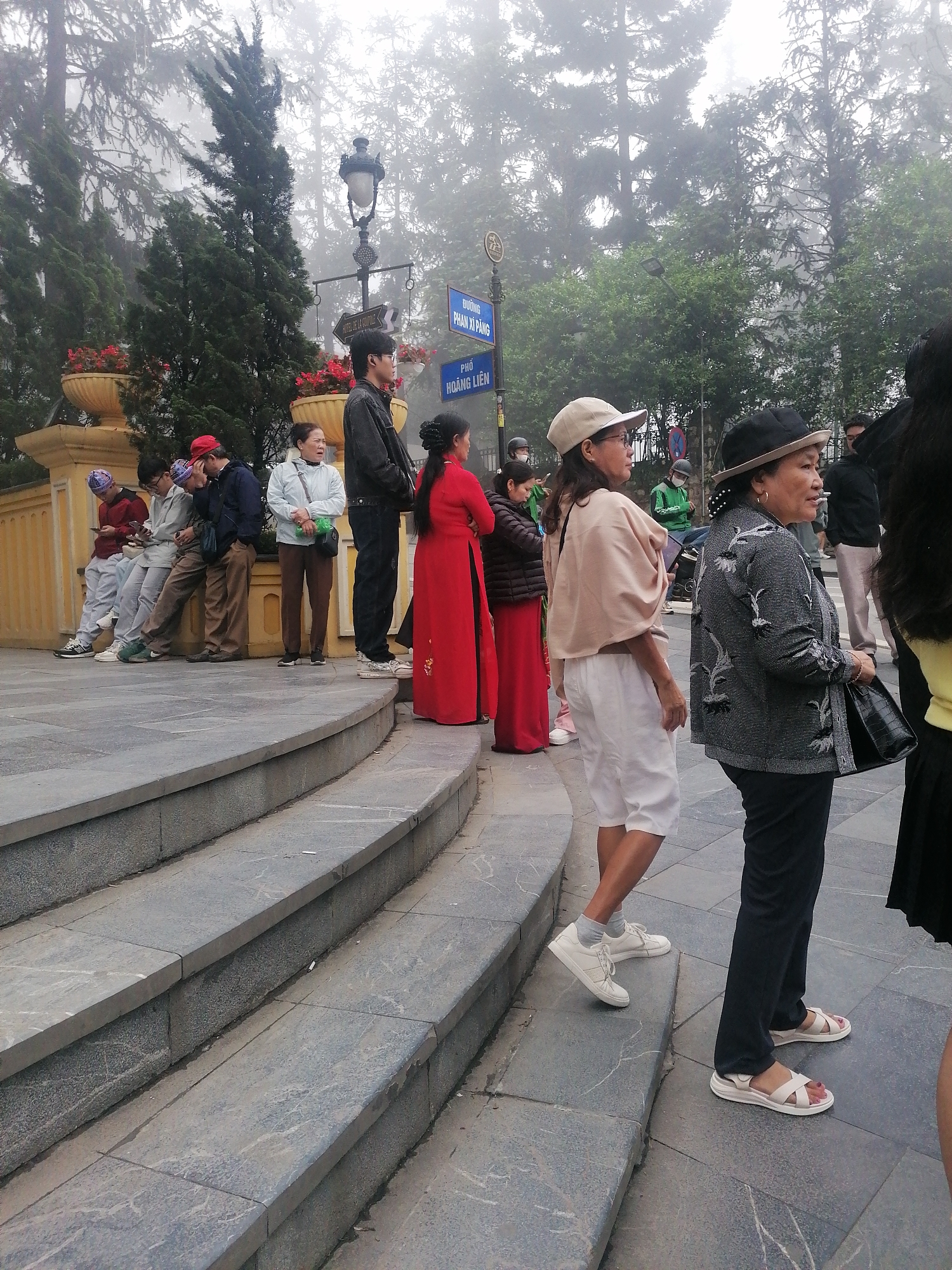 The image depicts a misty scene in Sa Pa Ward, Vietnam. A group of people stands and sits on and around a set of gray stone steps. The people are dressed in casual attire, with some wearing traditional Vietnamese clothing, such as the red ao dai. Several people appear to be tourists. The weather is foggy, with trees and a lamp post visible in the background. A street sign indicates "Phố Hoàng Liên" and "Đường Phan Xi Păng." The scene conveys a sense of a gathering or waiting.