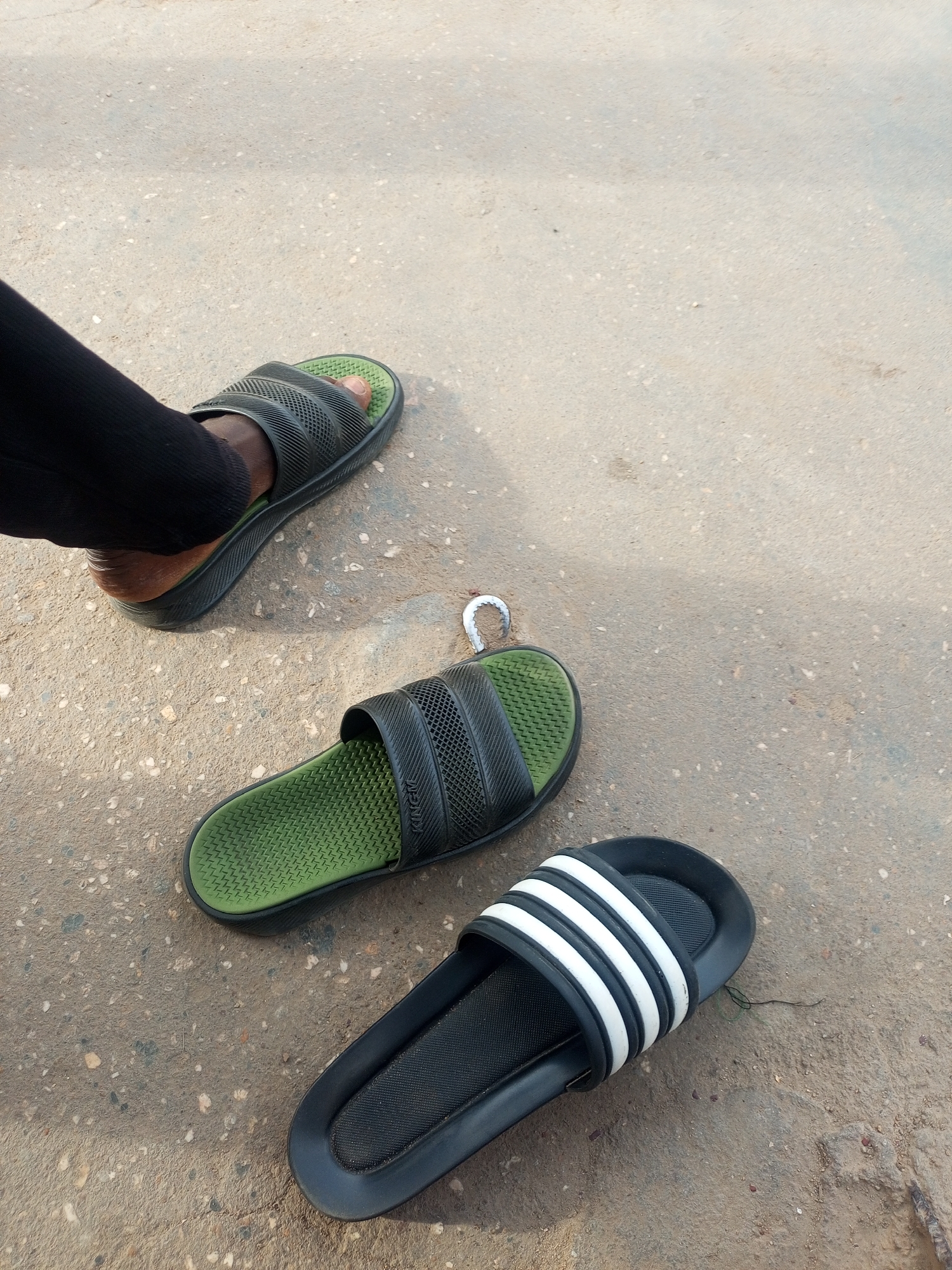 The image shows three sandals on a concrete surface, likely a street. One sandal is being worn by a person whose leg is visible, clad in black trousers. The sandals are of varying designs: one has three white stripes on a black background, and two are black with green insoles. The location is specified as Maiburiji, Nigeria, based on the prompt, suggesting the photo was taken there.