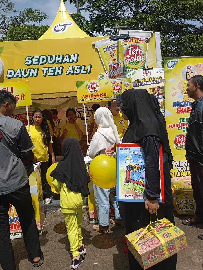 A group of people, primarily women and children, stand around a table in front of a brightly colored marketing display for "Teh Gelar" tea. The table features a display of tea products, with the brand name prominently displayed.  The women are wearing brightly colored clothing, including a young girl in a yellow outfit, a woman in a pink shirt and a woman wearing a black hijab.  There is also a man, visible in the background, wearing a white shirt and a black baseball cap. One woman is holding a drawing in her hand, while another is holding a large yellow balloon. The scene appears to be an outdoor event, likely a promotional booth for "Teh Gelar" tea. The location of the event is Soreang, Indonesia, based on the coordinates provided (latitude: -7.021, longitude: 107.53).