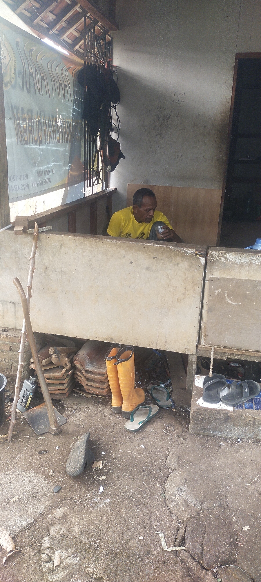 A man wearing a yellow t-shirt is sitting at a small counter. He is holding a clear glass in his right hand and appears to be drinking from it. The counter is made of concrete and wood. Behind the man, there is a wall with some writing on it. In front of the counter, on the ground, there are several objects, including a hoe, a stack of terracotta tiles, a pair of orange rubber boots, and several pairs of flip-flops. The environment appears to be an outdoor workspace, possibly a shop or repair stall, in Pasirhuni, Indonesia. The ground is dusty and there is some debris scattered around. Sunlight is streaming in from the top left of the image.