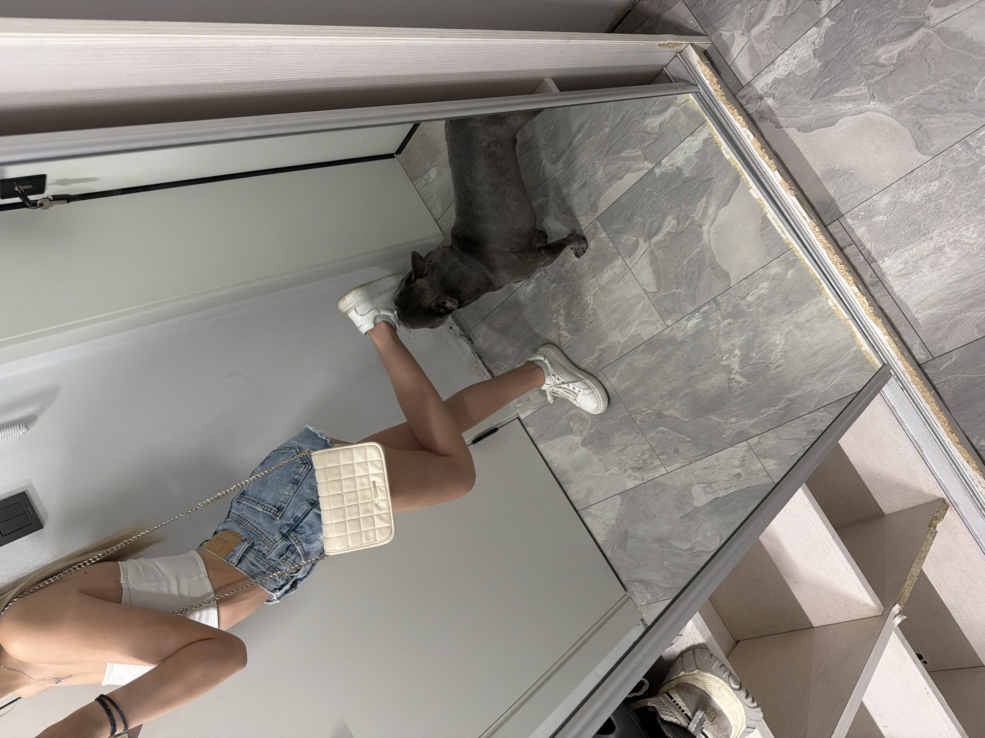 The image shows a young woman standing in front of a mirror, dressed in a white tube top, denim shorts, and white sneakers. She is accessorized with a white quilted purse and a delicate necklace. Her left leg is bent and extended towards the mirror. A grey French bulldog is standing on the floor in the reflection of the mirror, looking up at the woman's foot. The environment appears to be indoors, possibly a hallway or a room with a large mirror. The floor is tiled with a grey marble pattern. The background wall is white. The image is a reflection captured in a full-length mirror, which also shows a shelf unit to the left of the woman. There is a door or closet to the right of the mirror. The overall lighting is bright, suggesting an indoor setting during the day.