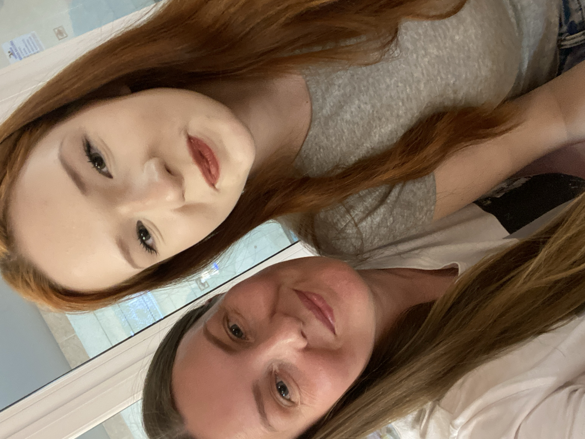 Two women are in the frame. The woman on the top has long, red hair and is wearing a grey t-shirt. She is looking at the camera and smiling with red lipstick. The woman on the bottom has long, light brown hair and is wearing a white t-shirt. She is looking at the camera with a slight smile. A window is visible in the background. It is not possible to confirm the location as the city of Chaikovsky, Russia, from the visual information provided.