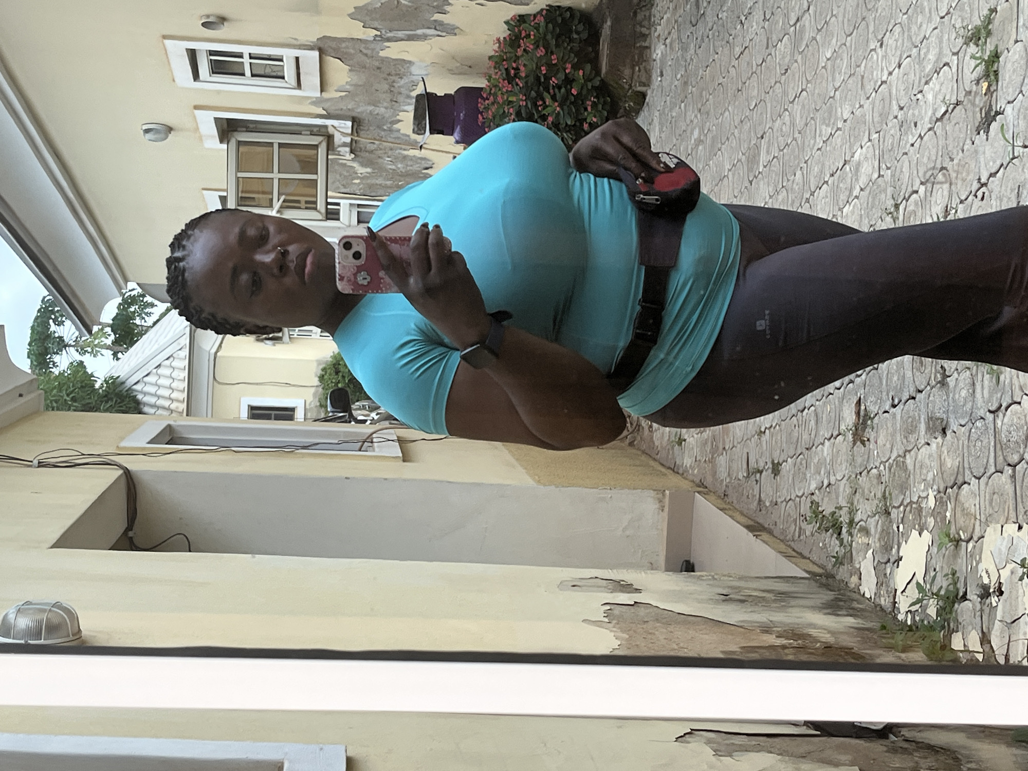 A full-body, slightly off-center, medium-shot, selfie photograph taken in an outdoor urban setting. A Black woman with braided hair and a nose ring is standing and holding a pink cell phone, looking at it. She is wearing a bright teal t-shirt and dark gray leggings. A black belt with a small, red and black pouch is around her waist. The woman is standing on a paved walkway made of light gray, circular, interlocking stones. The walkway is bordered on the left by a pale yellow building with peeling paint and on the right by more of the same building. In the background to the right, there is a large purple gas tank next to a bush with pink flowers. The building has several windows, some with shutters, and external light fixtures. There is some overgrowth of green plants and weeds in cracks in the paving and at the base of the building. The scene appears to be in Abuja, Nigeria.
