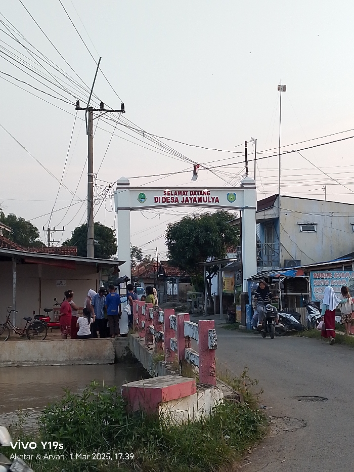 The image shows a street scene in the village of Desa Jayamulya, Kecamatan Haurgeulis, Indonesia. There is a sign that reads "Selamat Datang Di Desa Jayamulya." The sign is located on a small bridge over a waterway with green grass growing on the banks. A woman is riding a motorbike on the street. Several people are gathered on the left side of the image, near a small hut. The sky is clear and a few electric poles with wires run across the scene. There are several buildings in the background. It appears to be late afternoon or early evening.