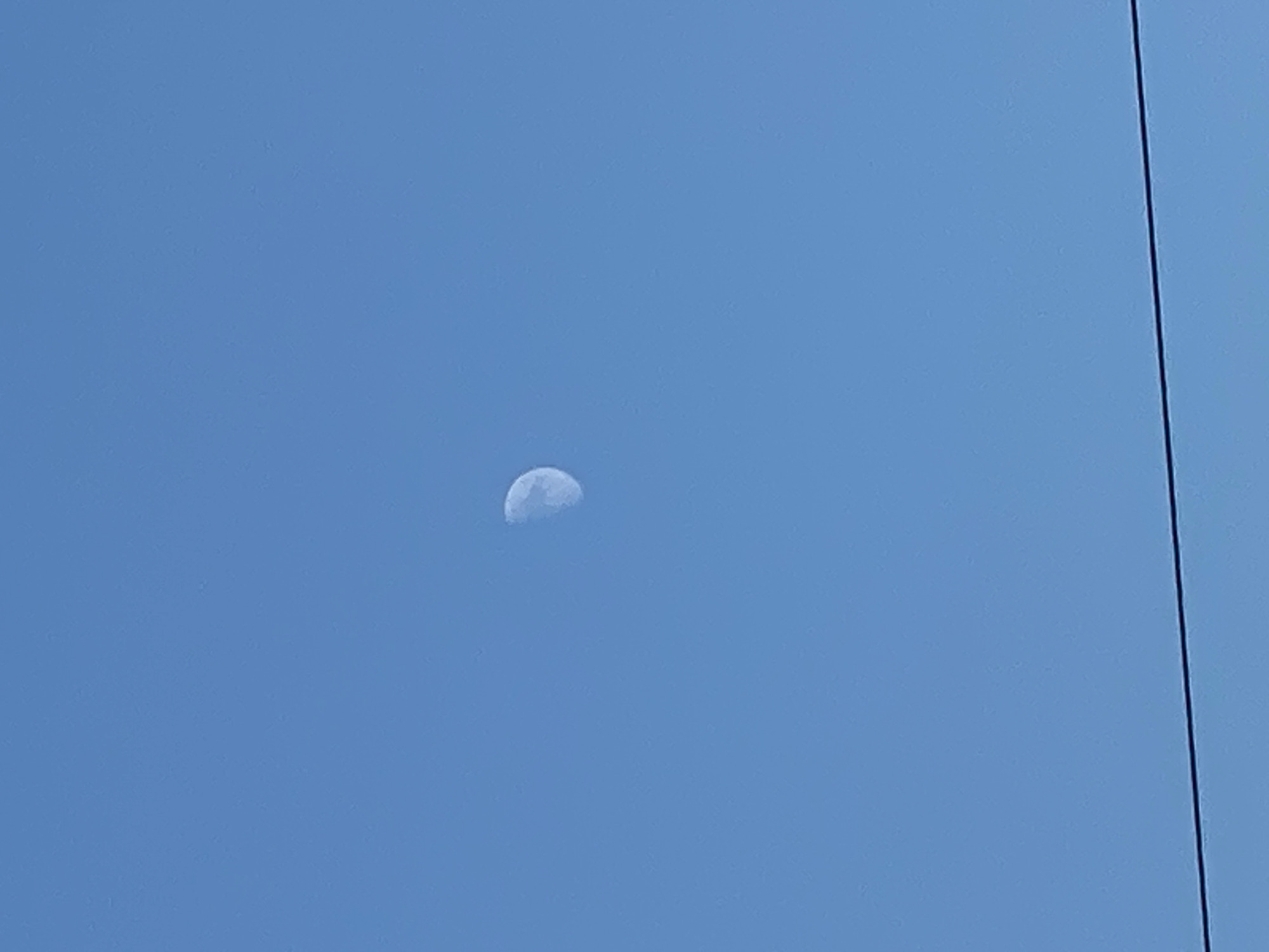 The image shows a clear blue sky with a crescent moon visible in the upper center. A dark horizontal line, possibly a wire or branch, runs across the lower portion of the image. The scene appears to be an outdoor view from Bjeliši, Montenegro. There are no people or specific landmarks visible that would indicate a precise location within the city.