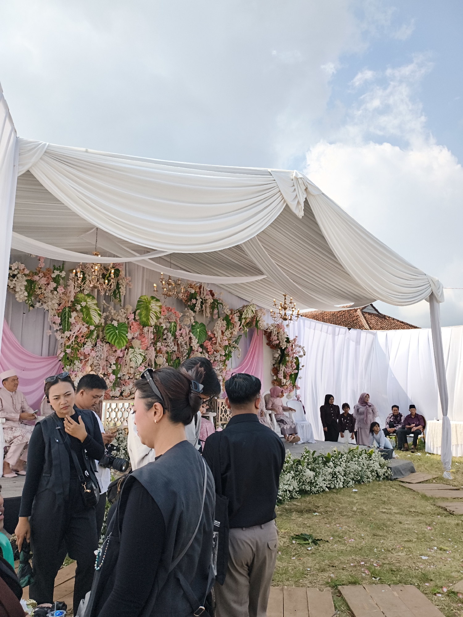 This outdoor scene captures a wedding event in Pacet, Indonesia. A white draped tent covers the wedding stage, which is decorated with an array of flowers and chandeliers. Several individuals are seated on the stage, including women in hijabs and men in traditional attire. In the foreground, several people stand in a cluster, some with cameras around their necks, suggesting they may be involved in event documentation. The ground is partially covered with grass and wooden planks. The background shows a building with a terracotta roof. The sky above is partly cloudy.