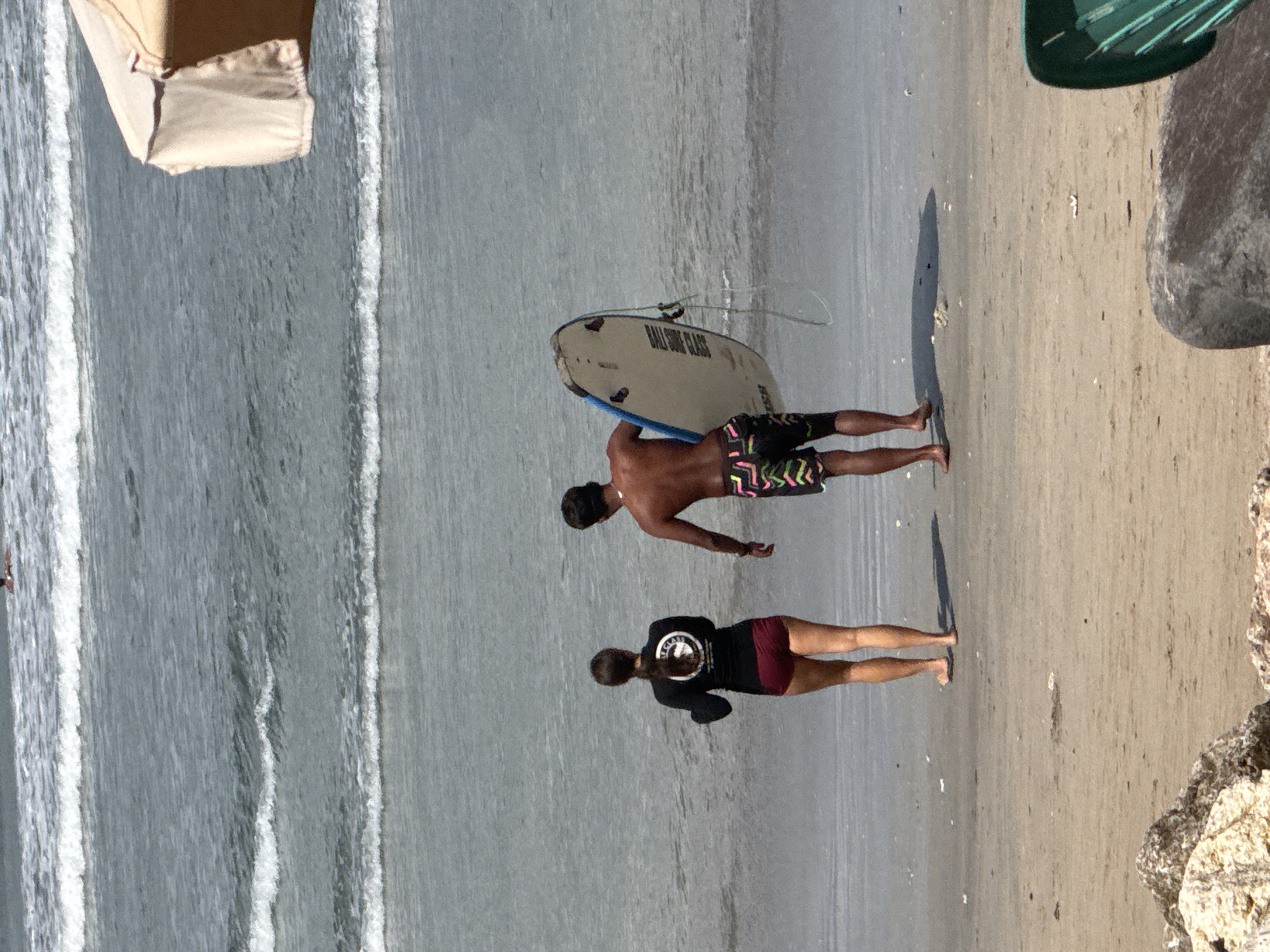 The image depicts a beach scene in Kuta, Indonesia, with two individuals at the water's edge. On the left, a person wearing a black rash guard and maroon shorts stands facing the ocean. To the right, a person in colorful swim trunks carries a surfboard. Gentle waves are breaking on the sandy shore, and a portion of a beach umbrella is visible in the upper right corner. The overall atmosphere suggests a typical day at a popular surfing destination.
