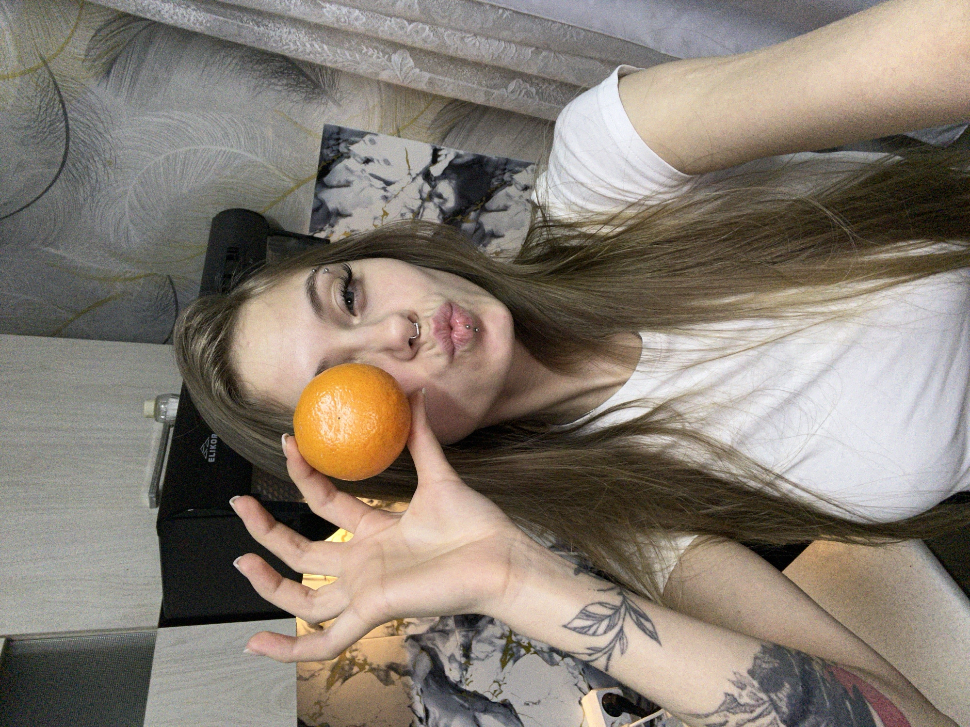 A young woman lies on her side, holding an orange in front of her face. She is wearing a white t-shirt and has long brown hair. She has tattoos on her left arm and piercings in her eyebrow, nose, and lip. The background includes a patterned wall, a marble-patterned surface, and a black object with the word "ELIKOR" on it. The photograph was not taken in the city of Фоки, Russia.