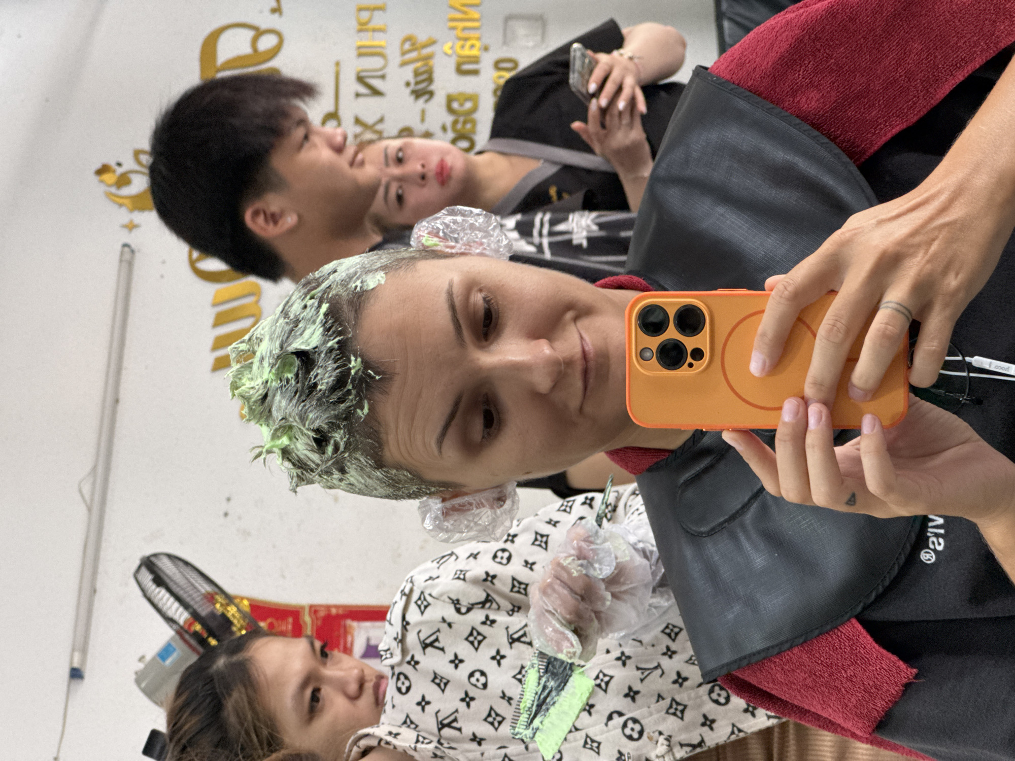 In a hair salon in Nha Trang Ward, Vietnam, a woman with short, light brown hair is taking a selfie in a mirror. Her hair is covered in a bright green hair dye, and she is wearing clear plastic coverings over her ears. She is dressed in a black smock with a red towel draped over her shoulders. In her right hand, she holds an orange smartphone, capturing the reflection of herself and the salon behind her.
To her left, another woman with dark hair, wearing a white jacket with a repeating monogram pattern, is applying hair dye to the back of the first woman's head with a comb. She wears clear plastic gloves. Behind them, a man with dark, short hair is visible, looking down. Further back, another woman in a black uniform holds a smartphone. The salon environment is suggested by the white walls, a fan, and a sign with gold lettering in the background.