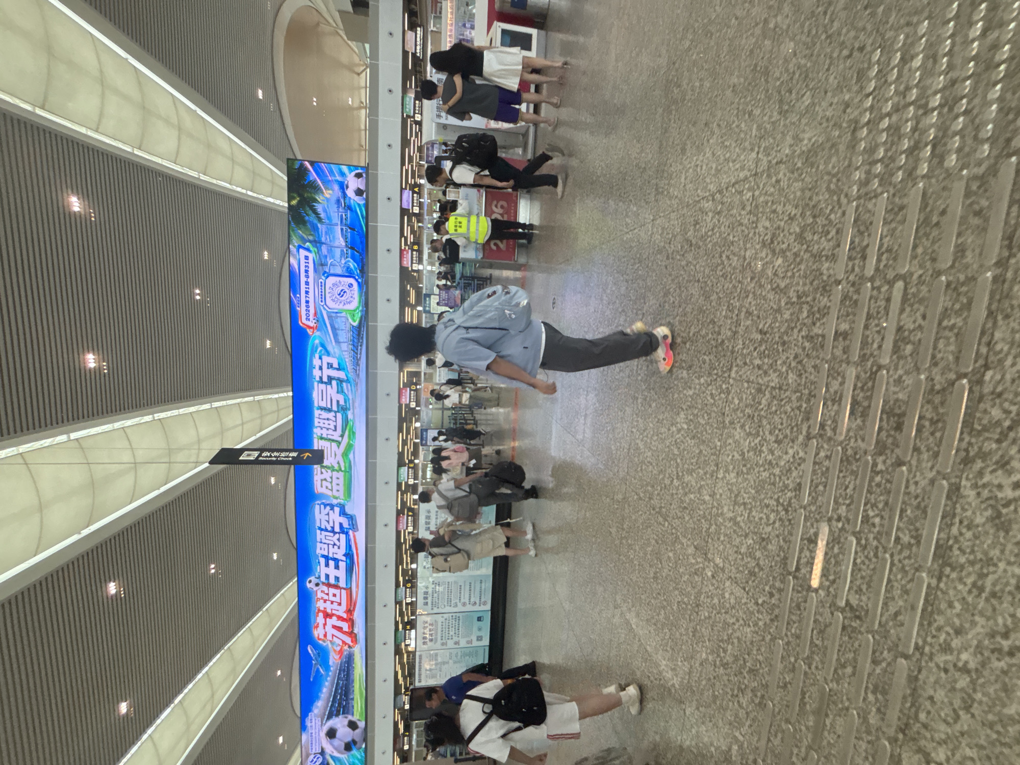 The image depicts an indoor public space, likely a transportation hub like a train station or airport, with a high, modern ceiling featuring prominent lighting fixtures and ventilation systems. A large digital billboard stretches across the upper portion of the scene, displaying promotional content with images of soccer and palm trees, and text in Chinese. Below the billboard, a row of service counters or ticket windows is visible, some with people interacting. The floor is made of speckled granite tiles, with tactile paving strips indicating a walkway.
Several individuals are present in the image, walking and standing. In the foreground, a person in a light blue shirt and dark pants with colorful shoes is walking away from the camera, carrying a large backpack. To the left, a person in a white t-shirt and shorts with a black backpack is walking towards the camera. Other people in the mid-ground are interacting at the service counters, queuing, or walking in different directions. One individual in a bright yellow vest appears to be a staff member or security personnel. The overall atmosphere suggests a busy, functional public area. The scene is set in the city of Xinwu District, China.