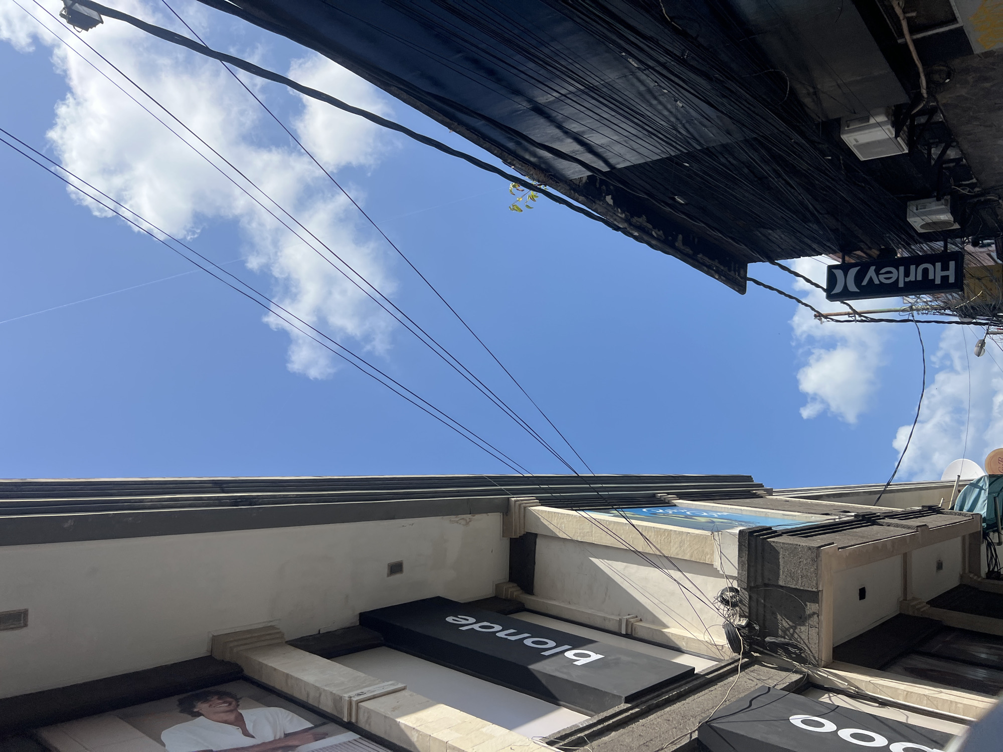 This is a ground-level, eye-level shot looking upwards at a bright blue sky filled with scattered white clouds. Two buildings frame the sky. On the left, a lighter colored building has a sign that reads "blonde" in white text on a black background. A portion of a large advertisement featuring a person in a white shirt is visible above the "blonde" sign. This building appears to have some architectural embellishments. On the right, a darker building, possibly made of wood or painted a dark color, is densely covered with black electrical wires. A sign reading "Hurley" with its distinctive logo is attached to this dark building. Two white air conditioning units are also mounted on the facade of the dark building. The composition suggests an urban environment in Kuta, Indonesia, characterized by narrow streets and visible infrastructure like utility wires.
