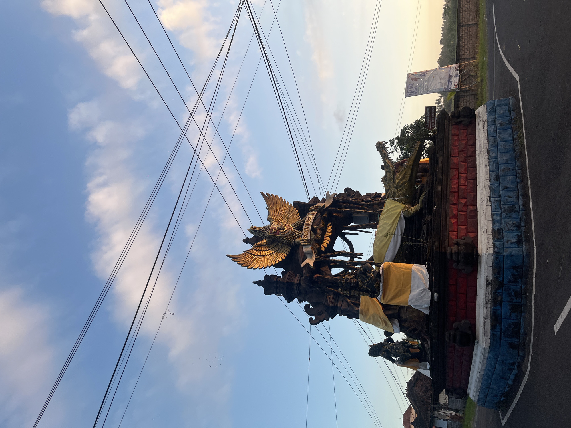The image depicts a monument in the city of Mendoyo, Indonesia, under a bright blue sky with scattered clouds and a network of overhead power lines. The monument is a multi-tiered structure with intricate carvings, prominently featuring a large golden eagle with outstretched wings at its apex, bearing the inscription "BHINNEKA TUNGGAL IKA". Below the eagle, various figures and mythological creatures are depicted, including a fierce-looking crocodile with its mouth agape. The monument's base is adorned with red and blue colored blocks. Yellow and white cloths are draped around parts of the structure, possibly as offerings or decoration. To the left of the monument, traditional Indonesian houses can be seen. To the right, a large banner is displayed on a wooden stand, and a brick wall lines the background. The monument is situated at a road intersection, with asphalt roads visible in the foreground. The overall scene suggests a cultural or religious landmark in a semi-rural setting.