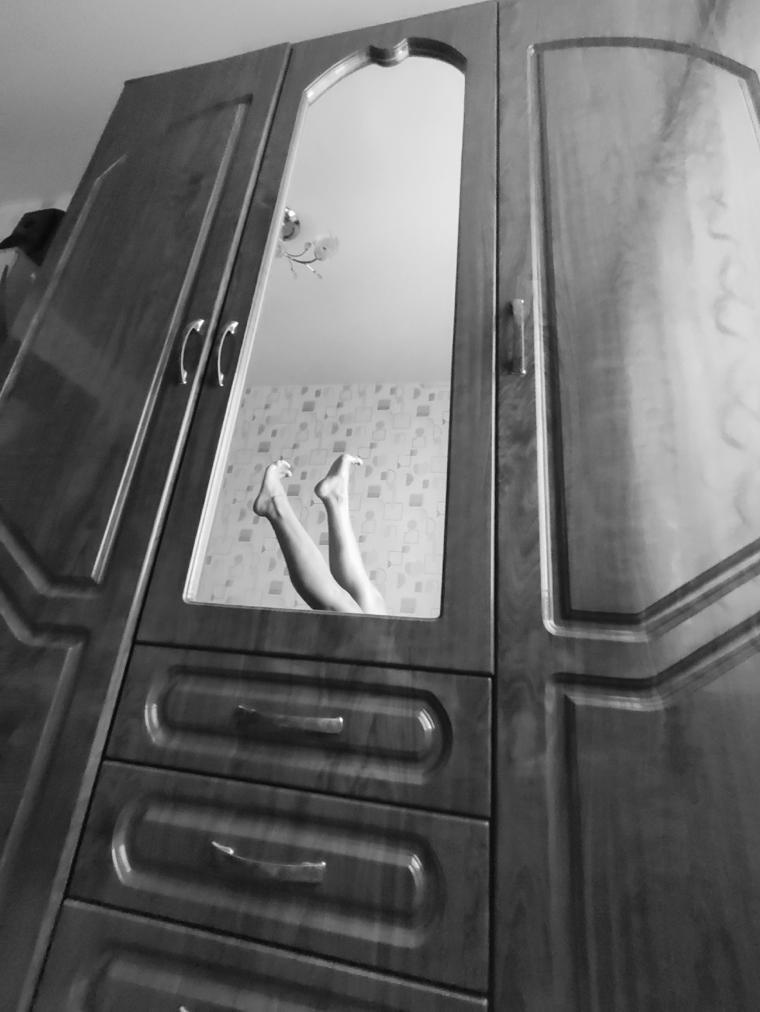 A black and white, low-angle shot depicts a wooden wardrobe with a mirror in the center door. Reflected in the mirror are a pair of human legs pointing upwards in a room with patterned wallpaper. A ceiling light fixture is also visible in the reflection. The wardrobe has two doors with handles and drawer pulls on the lower part. The location is presumed to be Chaikovsky, Russia.