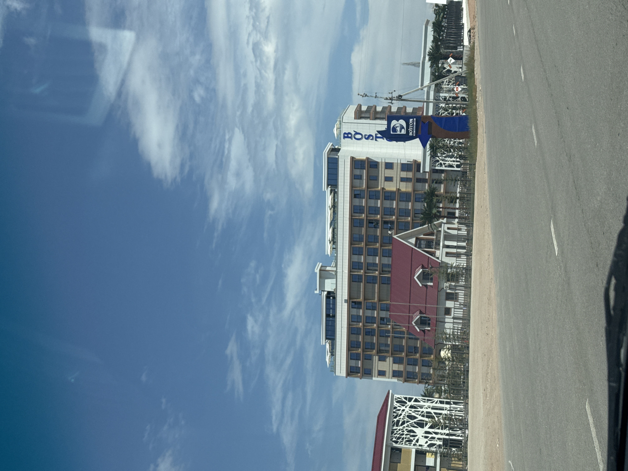 The image shows a modern building complex under a bright blue sky with scattered clouds. The main structure is a multi-story hotel or apartment building with numerous dark-paned windows and a white upper level featuring lounge chairs and umbrellas. To the left, a smaller building with a red roof has an abstract white decorative facade. In front of the main building, there is a shorter building with a red roof and dormer windows, connected to the larger complex. A prominent blue sign with the word "BOSTON" in large blue letters and a stylized "B" logo is positioned in front of the main building, indicating a commercial establishment. The area appears to be a developing or newly constructed area, with unpaved ground in the foreground and a paved road. The overall scene suggests a sunny day in a semi-urban or resort-like setting.