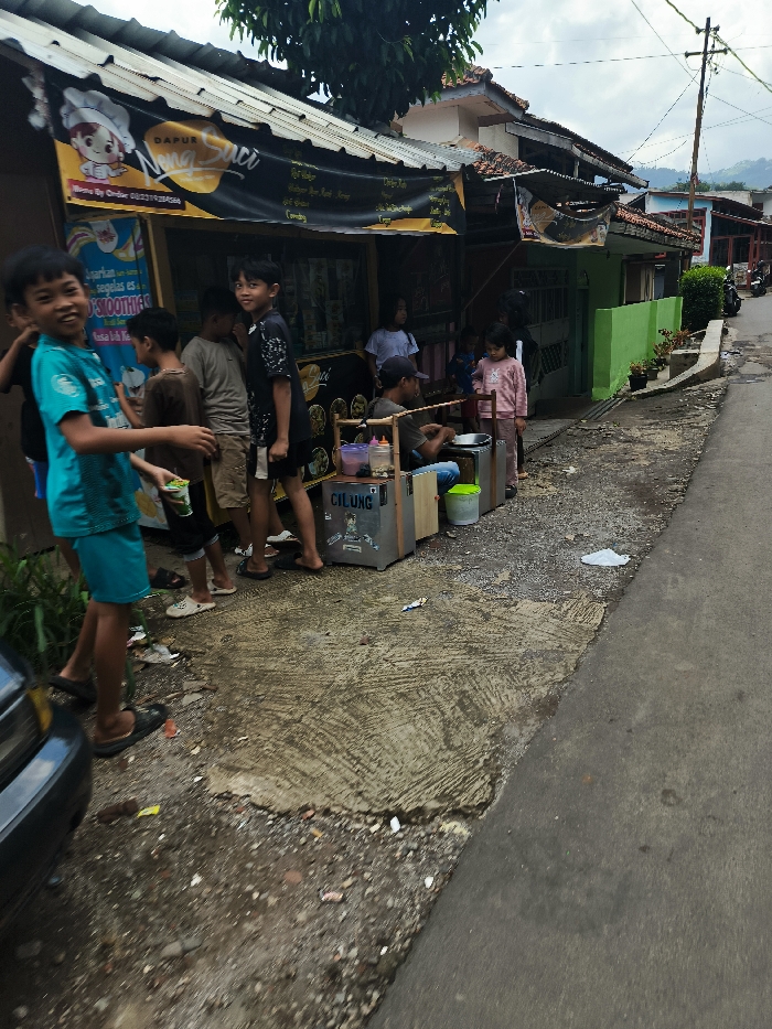 The image shows a street scene in Cimenyan, Indonesia, located at coordinates -6.885, 107.654. A group of people are gathered in front of a small storefront with a sign that reads "Dapur Saci". There are several men and women standing around a small table where a man is seated, apparently preparing food. The storefront is adorned with a yellow awning and has a sign advertising "smoothies" on the wall. There are a few potted plants on a small wall behind the group, and an alleyway leads to the left, toward a paved street. The pavement is cracked and worn. To the right of the storefront, there is a small house with a green wall. A motorcycle can be seen in the background, and there are several power lines in the sky. The people are standing in a line, some of them looking towards the camera. The person in the forefront is wearing a blue and green t-shirt and shorts. They are standing with their feet on the ground and their hands in front of them. The people in the background are blurred, but they appear to be moving around the table and the storefront. The scene appears to be a typical street scene in a small town in Indonesia.