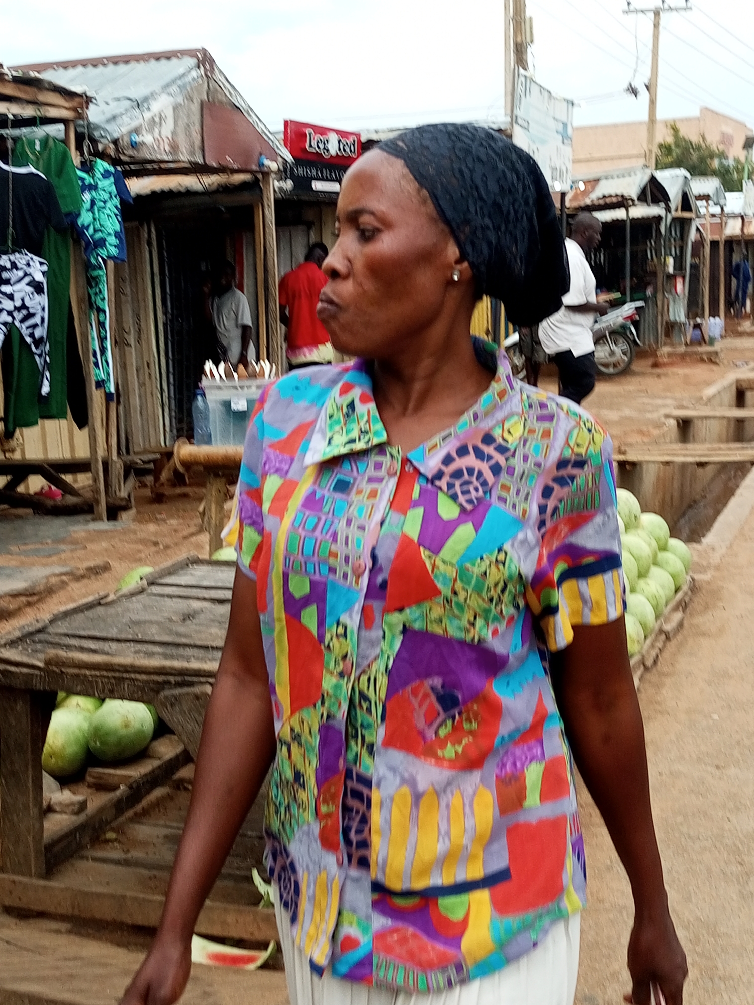 The media file shows an outdoor scene in a marketplace in Gombe, Nigeria. The primary subject is a woman in the foreground, facing to her right and looking slightly away from the camera. She is wearing a vibrant, multi-colored patterned short-sleeved shirt and a black headscarf. Her expression is neutral, and she appears to be standing.
Behind the woman, the setting reveals a bustling marketplace with various stalls. One stall displays clothing, including green and black items hanging on a rack. Further back, another stall has a sign with red lettering that appears to read "Legted" and "SHISHA FLAVOR". Several people are visible in the background, including a man in a red shirt and another in a white shirt near a motorcycle.
The ground is dusty and appears to be a dirt path. To the right of the woman, there is a display of round, green fruits, possibly gourds or melons, stacked in a wooden crate. Wooden structures and tables are scattered throughout the scene, indicative of market stalls.
The time of day appears to be daytime, given the lighting, though the sky is overcast, suggesting potentially cloudy weather. The overall impression is that of a lively, everyday market environment. There is no specific activity or event clearly depicted beyond general market operations.