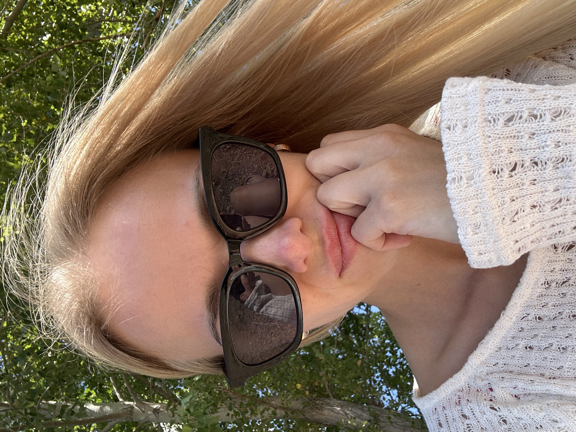 A young woman with long blonde hair is captured in a close-up selfie. She is wearing large, dark-framed sunglasses and a white, open-knit sweater. Her right hand is resting on her chin, and her lips are slightly pursed. The background shows green foliage and tree trunks, suggesting an outdoor setting. There are no discernible urban elements to suggest a specific city location like Gornyi Erik, Russia.