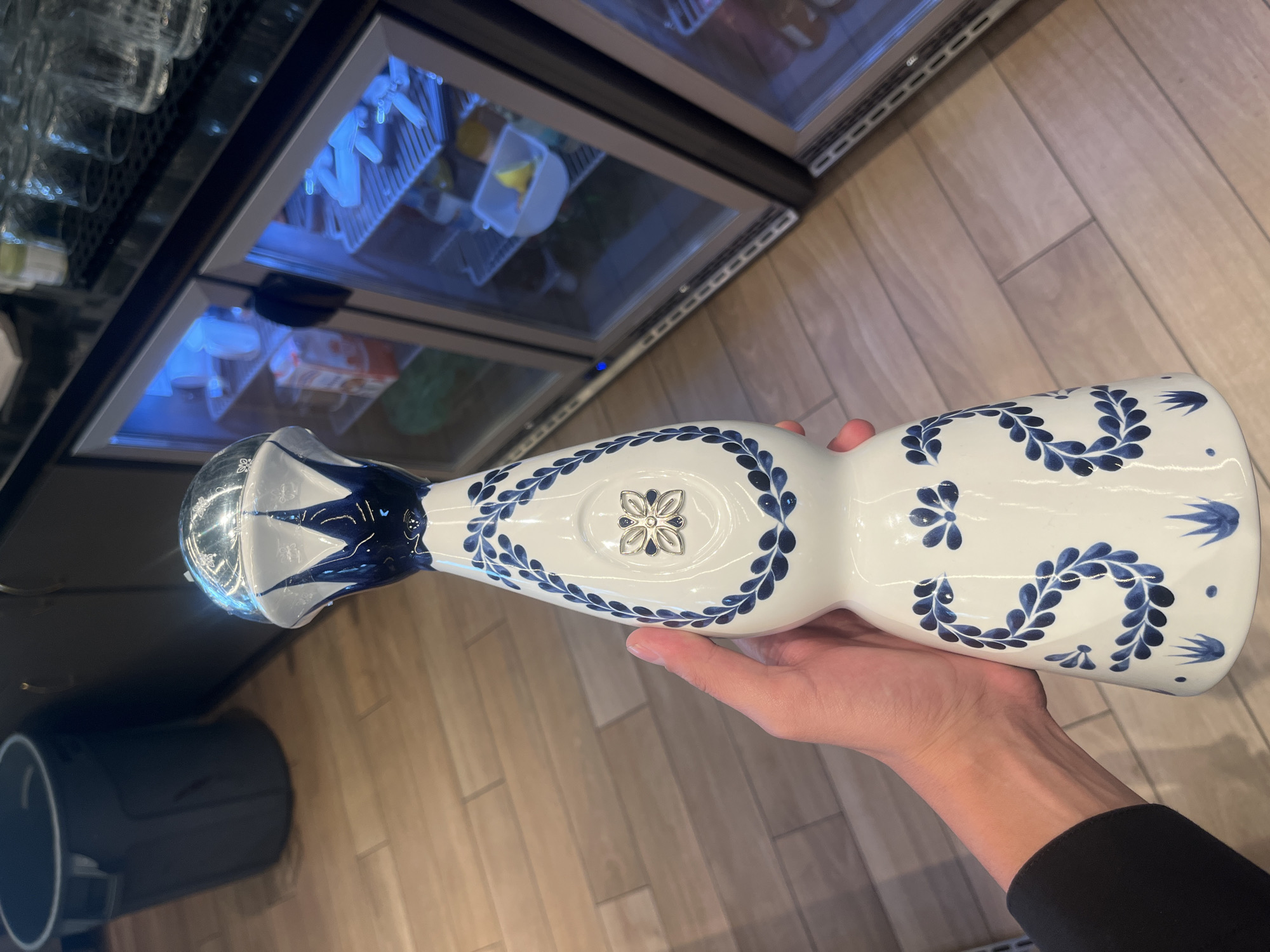 A close-up, horizontally oriented shot captures a person's hand holding a distinctively decorated bottle. The central object is a white ceramic or porcelain bottle, elongated and tapering, adorned with intricate blue floral and foliage patterns. A silver-toned, four-petal flower motif is embedded on one side of the bottle's body. The bottle's short, bulbous neck is topped with a clear, possibly glass or crystal, stopper or cap, which also features blue and white patterned details.
A person's right hand, with fingers and thumb visible, supports the bottle from underneath. The individual wears a black long-sleeved garment on their forearm.
In the background, on the left, several clear glass cups are visible on shelves. Behind and above these, two glass-fronted display refrigerators or beverage coolers extend across the frame. Their interiors are illuminated with a cool blue light, revealing indistinct items stored within. The floor, visible on the right, is made of light-colored wooden planks.
The setting appears to be an indoor commercial space, such as a bar, restaurant, or retail store, in Sofia, Bulgaria. The lighting is artificial, suggesting no particular time of day. No visible text is present in the image.