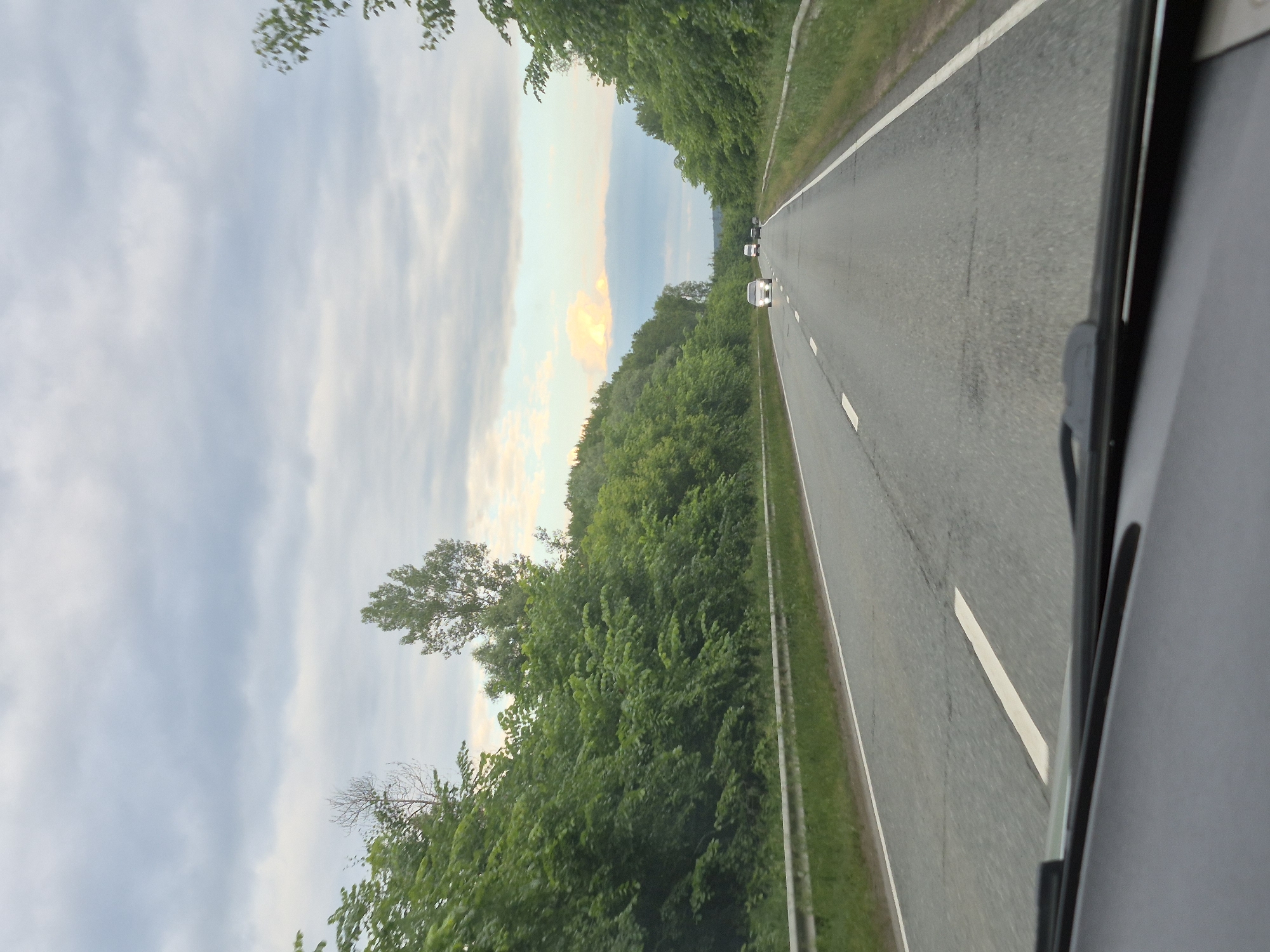 The image depicts a view from inside a vehicle traveling on a two-lane highway lined with dense green trees and vegetation. The road surface is asphalt with white lane markings. In the distance, several vehicles are visible on the road, including a white van and at least two cars. The sky is overcast with clouds, but there are hints of lighter sky and possibly a sunset or sunrise in the far distance, with a patch of light orange and blue clouds. The scene appears to be a rural or semi-rural setting, possibly on the outskirts of Niva, Russia. The perspective is from the front passenger seat or driver's view, looking through the windshield, which is partially visible at the bottom of the frame.