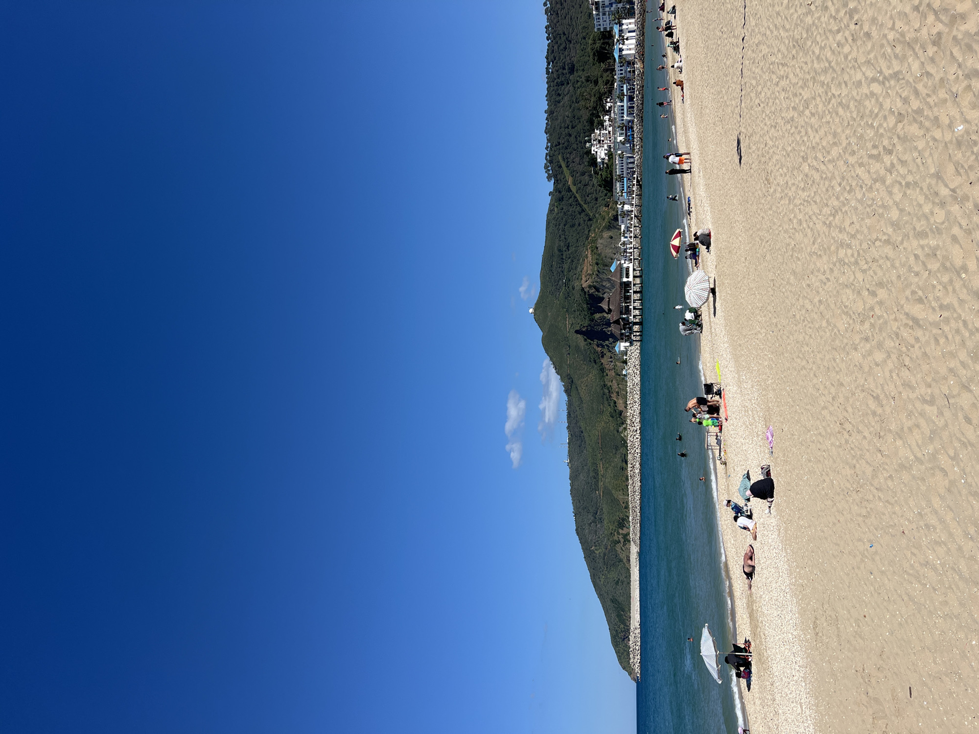 The image shows a beach in M'Diq, Morocco. To the left is a clear blue sky, occupying about half of the frame. The right half depicts a beach with light brown sand. Along the shoreline, there are people sunbathing, swimming, and relaxing under umbrellas. In the background, a green, tree-covered mountain slopes down to meet the beach. Buildings and infrastructure are visible along the mountain's base. The water is a greenish-blue color. The beach appears to be a popular spot for recreation and leisure.