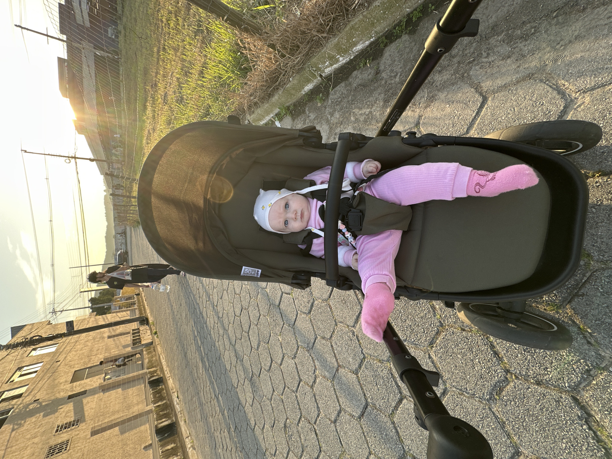 The image shows a baby sitting in a stroller on a cobblestone street in Florianópolis, Brazil. The baby is wearing a pink outfit and a white hat with a floral pattern. The stroller is dark green. In the background, there is a man walking with a bottle of water and a plastic bag. There are buildings and a fence on the side of the street. The sun is setting, casting a warm glow on the scene.