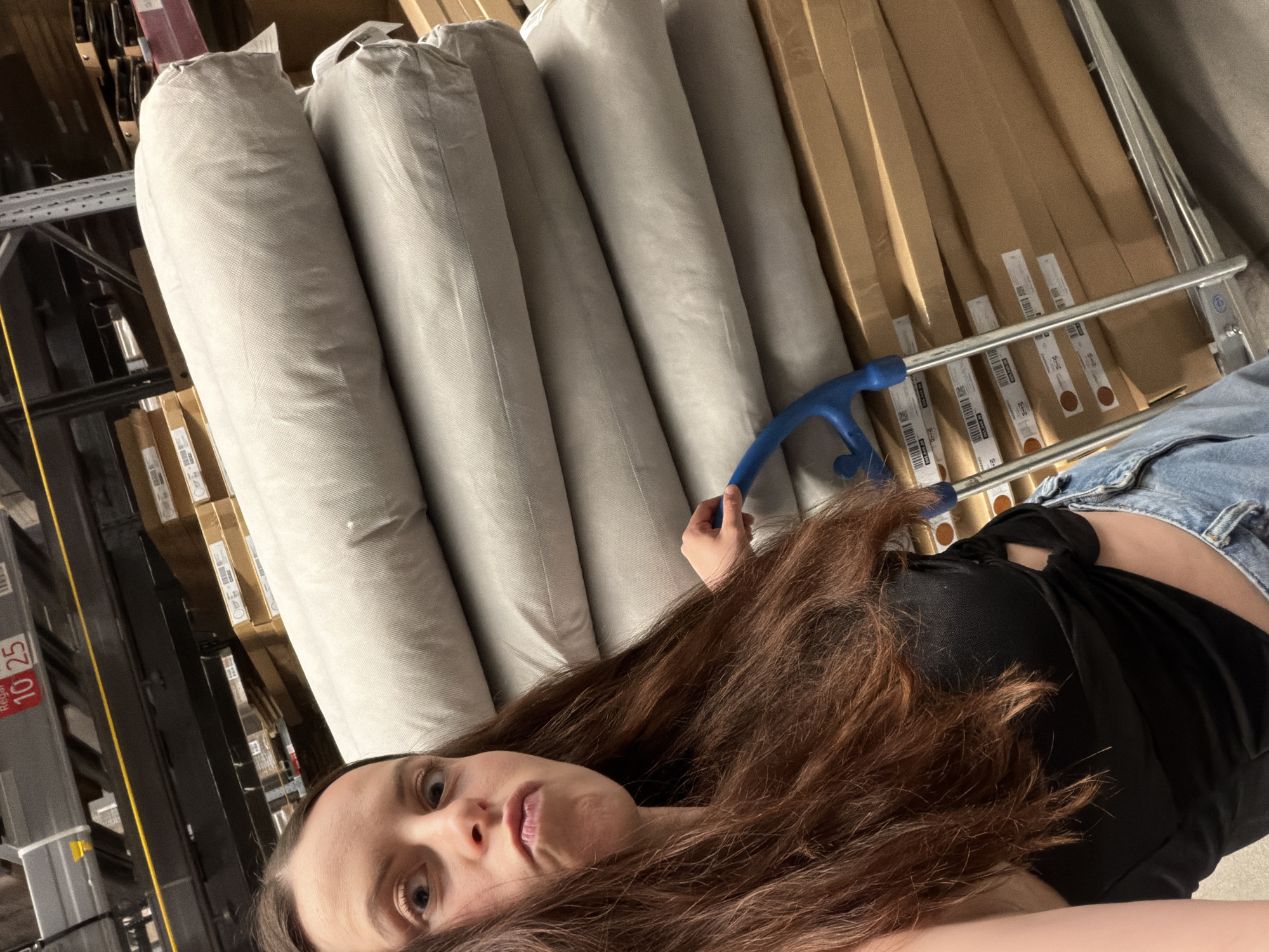 A person with long brown hair, wearing a black top and blue jeans, is in what appears to be a retail or warehouse setting. They are holding the handle of a blue cart containing rolls of fabric or material, and cardboard boxes. The location is possibly a store in Kaiserslautern, Germany.
