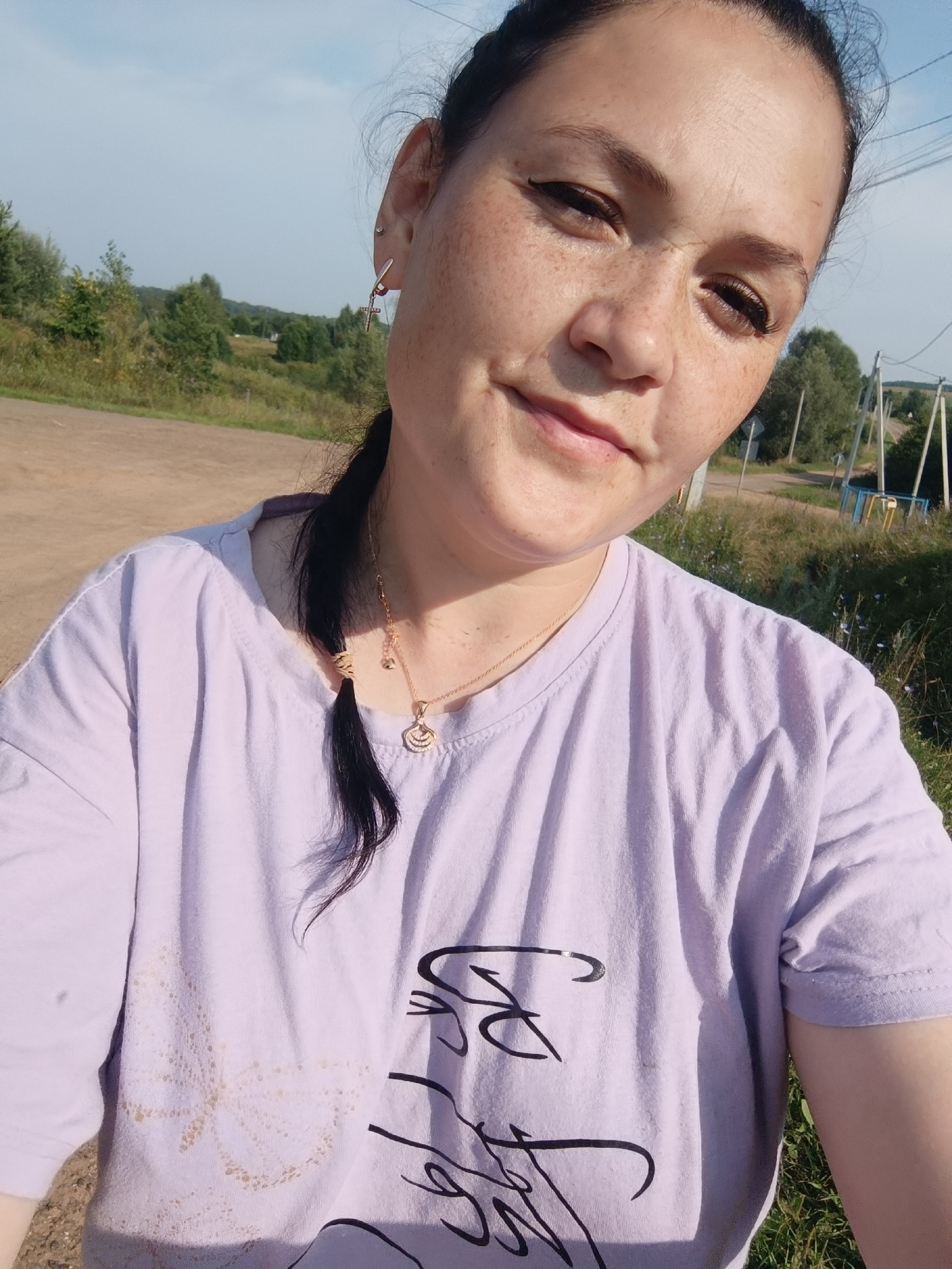 A young woman with dark hair pulled back in a ponytail, wearing a light purple t-shirt and a gold necklace, is posing for a selfie outdoors. She has freckles on her face and is wearing dangling earrings. The background shows a dirt road, trees, and a clear sky, suggesting a rural or suburban setting. There are no specific landmarks that identify this location as Karsha, Russia.