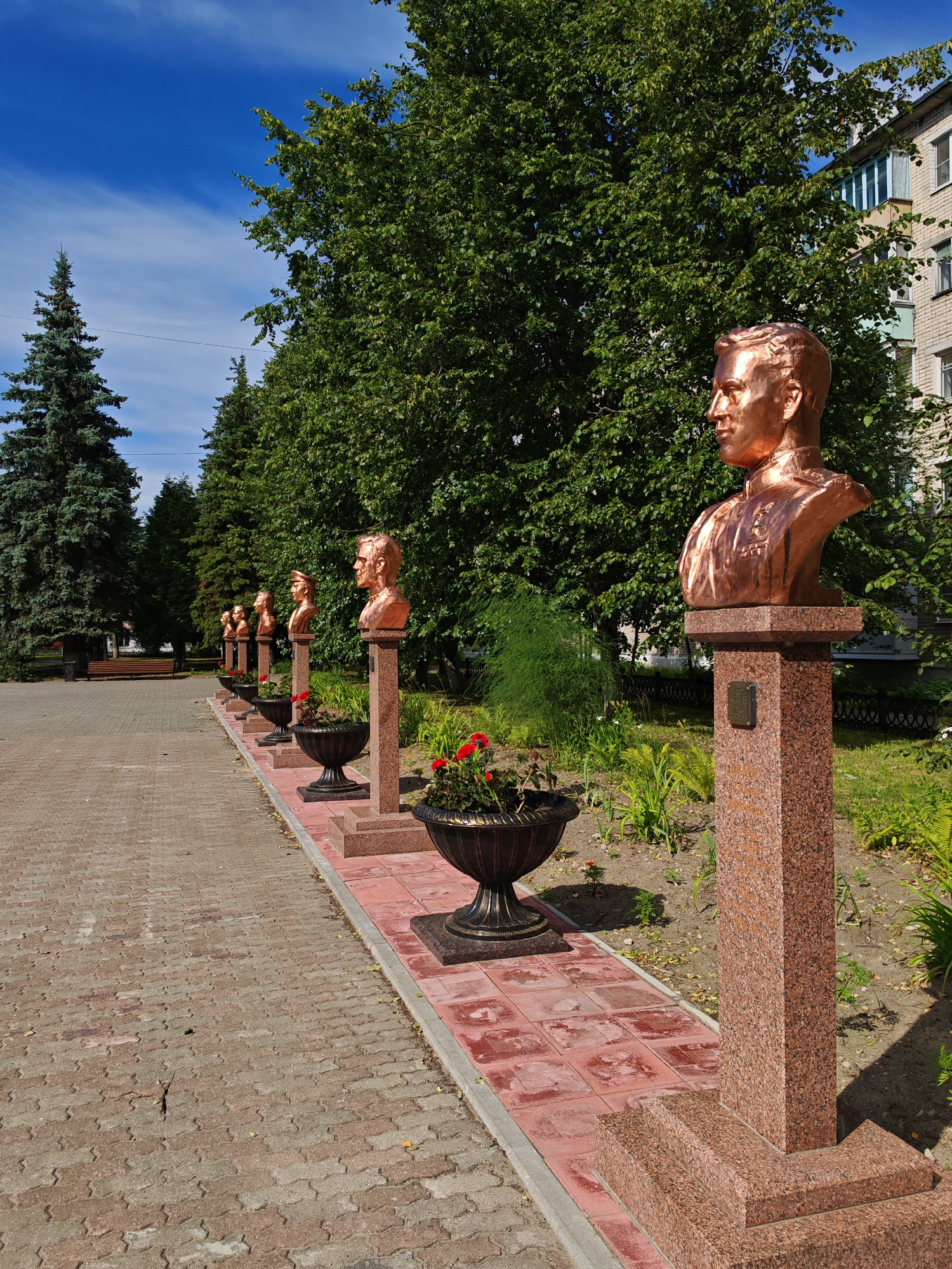 A paved pathway lined with red paving stones leads to a series of bronze busts on granite pedestals, positioned in a row against a backdrop of lush green trees and a brick building. The busts, facing to the left, are of men in military-style attire, each atop its own individual pedestal. The first pedestal, closest to the viewer, features a bronze bust of a man with medals on his chest, standing on a multi-tiered granite base. To the right of this pedestal, a dark, ornate planter holds red flowers. The pathway is flanked by a border of red tiles and a gray curb. Beyond the trees, a multi-story brick building with balconies is visible. The sky is a clear blue with a few wispy white clouds. This scene is located in the city of Buy, Russia, likely in a park or public square.