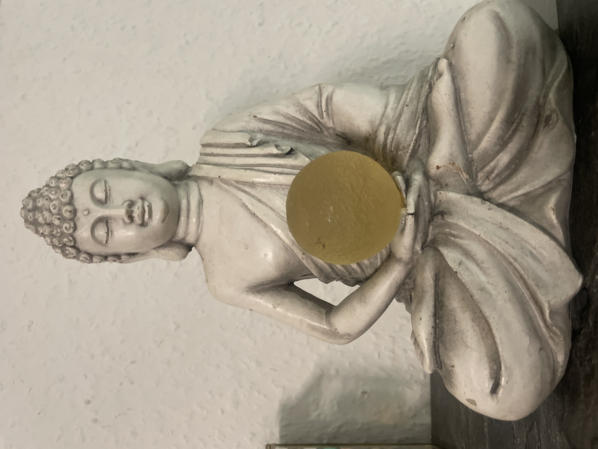 A white, seated Buddha statue is displayed against a textured white wall in Iserlohn, Germany. The statue is holding a translucent, yellow, spherical object in its hands, which are clasped together in its lap. The statue is detailed, with an intricately carved headdress and robe. The overall lighting suggests an indoor setting, possibly a home or shop.