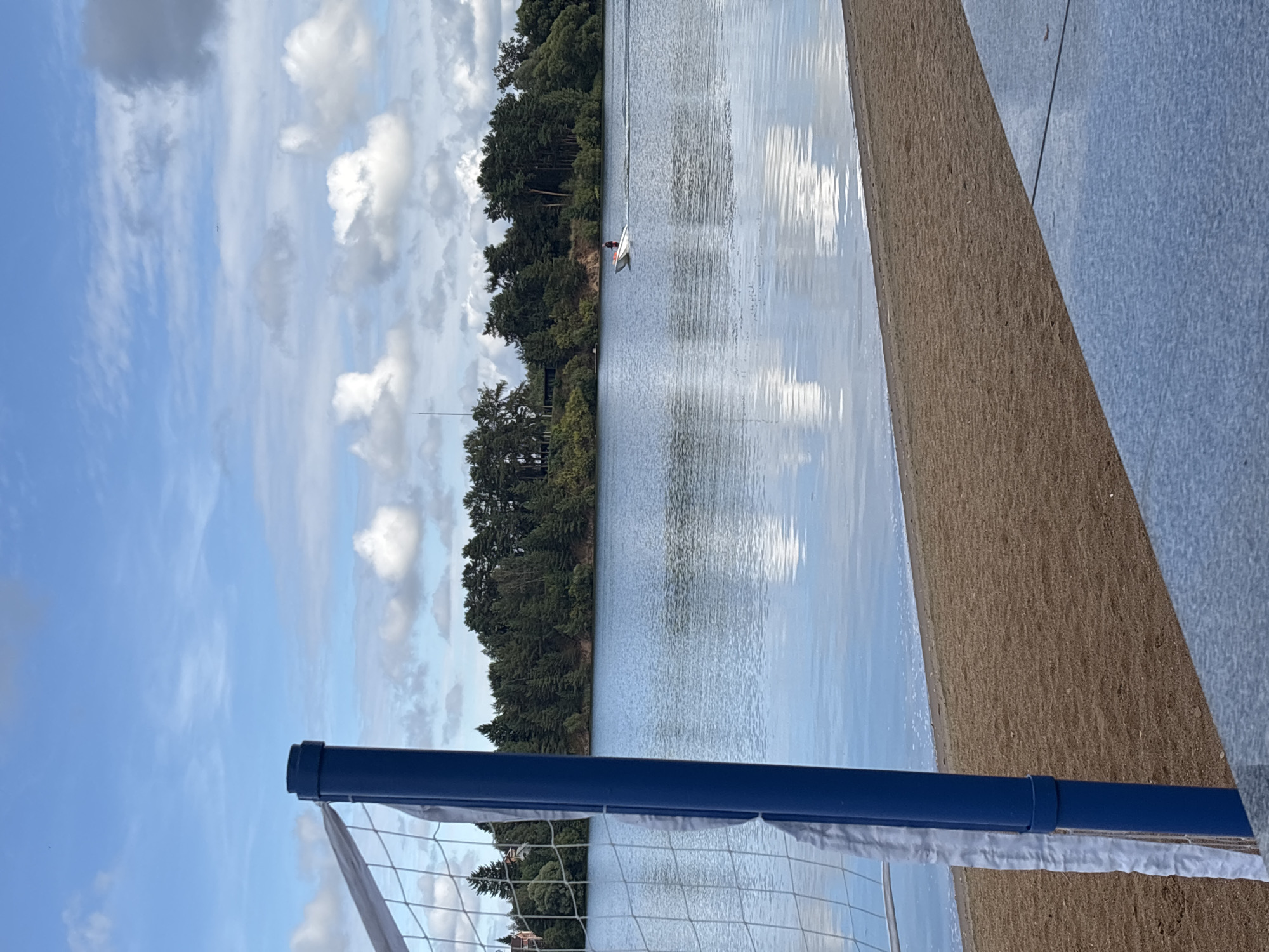 This is a horizontal, eye-level shot of a lake with a wooded shoreline and a blue sky with scattered clouds. In the foreground, on the left, is a blue pole with a white net, suggesting a volleyball court near the beach. The beach itself is made of sand, with a paved area in the lower right corner. On the lake, a small boat with a person in it is moving from left to right. The water is calm and reflects the sky and trees. The location appears to be a recreational area in the city of Рождествено, Russia, likely a park or a beach on a lake.