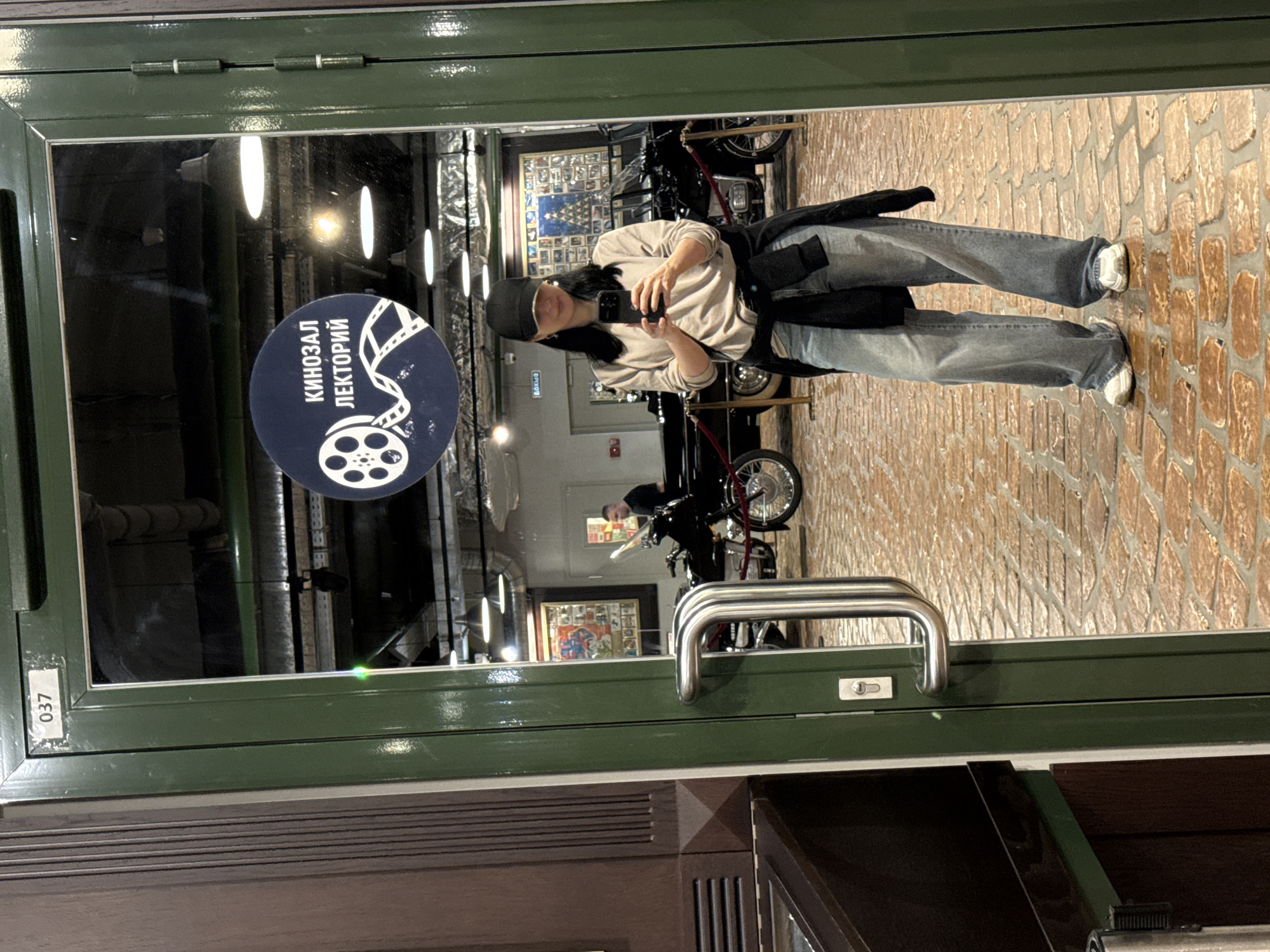A woman in a light-colored long-sleeved shirt, baggy denim jeans, a black baseball cap, and white sneakers is reflected in a glass door. She is holding a black smartphone, taking a selfie. A black jacket is tied around her waist. The background of the reflection shows a man in a dark shirt standing near a motorcycle. Another motorcycle and a black car are also visible. A circular sign with a film reel and Russian text reading "КИНОЗАЛ ЛЕКТОРИЙ" (Cinema Lecture Hall) is affixed to the glass. The floor is paved with reddish-brown bricks. The door is dark green with a silver handle. Above the door, a white sticker with the number "037" is visible. The environment appears to be a museum or exhibition hall, possibly in Arkhangel'skoe, Russia, given the text on the sign.