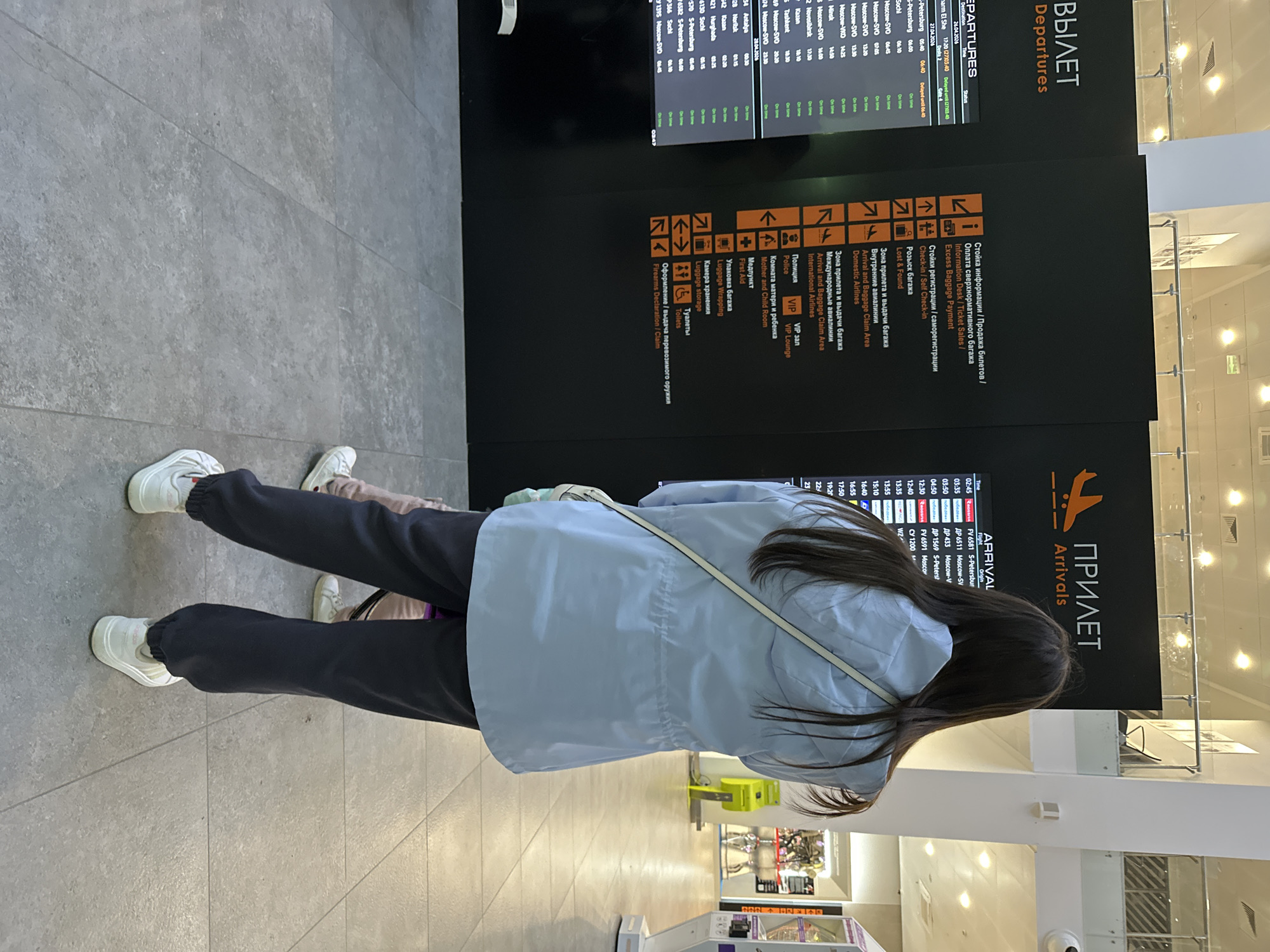 The image shows a woman standing in an airport terminal. She is looking at a flight information display board that shows flight arrival and departure information in both Russian and English. The board lists flights to and from various destinations. The terminal has a tiled floor and bright lighting. There are other people visible in the background and nearby shops. The text indicates that the airport provides information and services in both languages, including information desks, baggage claim areas, police and VIP lounges, a mother and child room, first aid, luggage wrapping, and firearms declaration services.