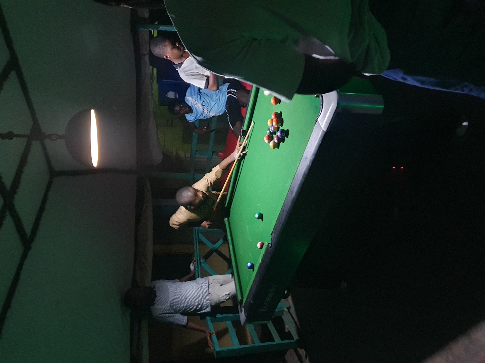 A group of young men are playing pool at an outdoor pool hall in Lokoja, Nigeria. The pool table is green felt with white pockets and the balls are scattered on the table. One of the men is about to take a shot, while the others are watching or waiting for their turn. The pool hall appears to be covered by a roof, and there are lights hanging from the ceiling. The overall atmosphere is casual and friendly.