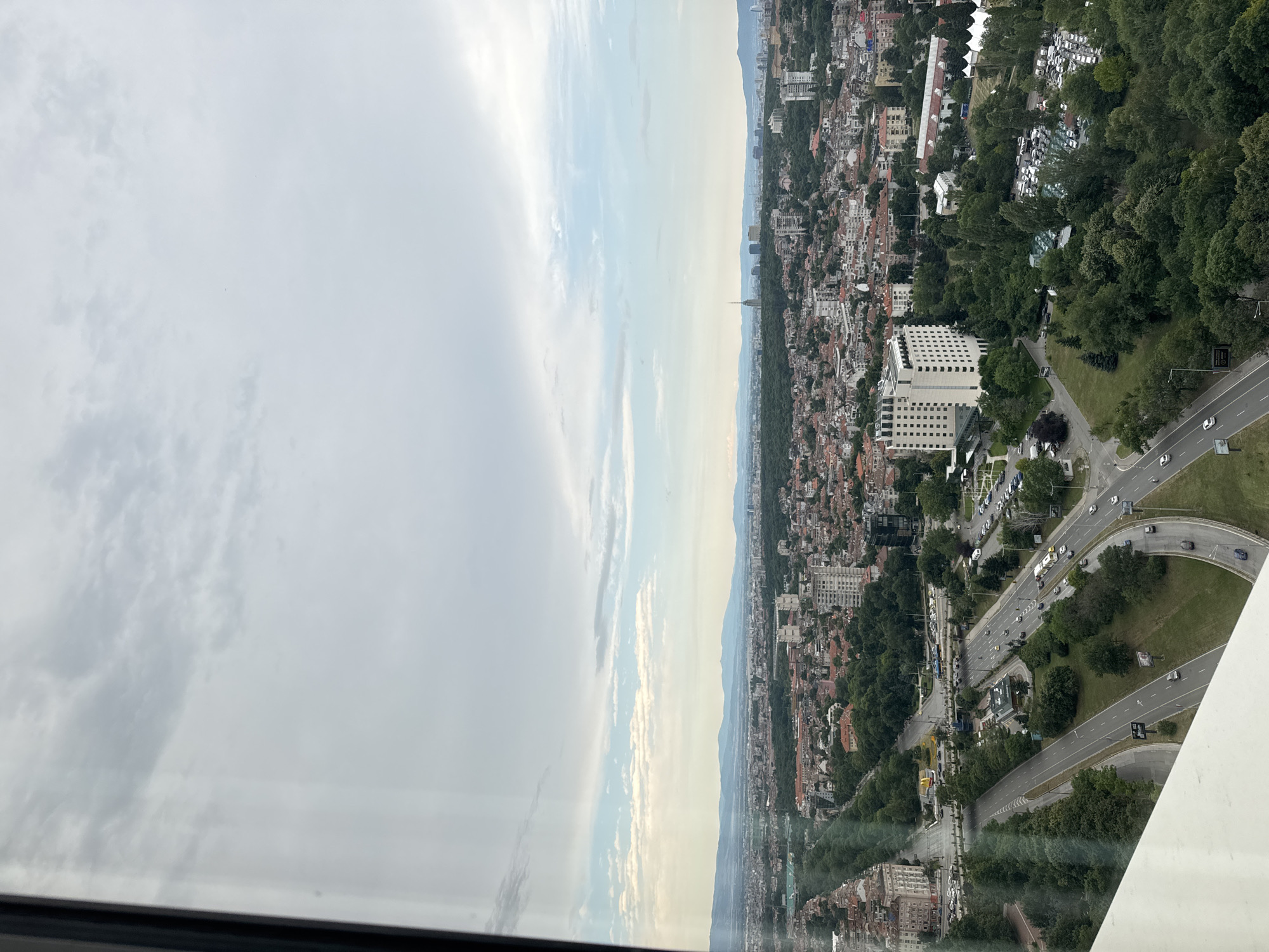 The image depicts an aerial view of Sofia, Bulgaria, taken from a high vantage point, likely a building. The scene overlooks a dense urban landscape characterized by a mix of residential and commercial buildings, interspersed with numerous trees and green spaces. A prominent feature is a wide, multi-lane road that curves through the city, with vehicles visible on it. In the mid-ground, a large, modern, white building with many windows stands out, surrounded by trees. Further in the distance, a hazy skyline is visible, with mountains forming a backdrop against a cloudy sky. The overall impression is of a sprawling city with a significant amount of greenery.