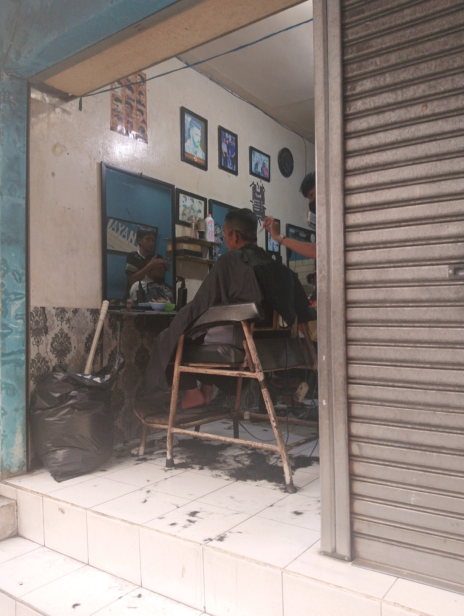This is a photograph of a barbershop. The shop appears to be located on a street in the city of Bandung, Indonesia. The shop is open, with a metal roll-up door partially open to reveal the interior. Inside, there are two barbers and one customer. One barber is reflected in a mirror, attending to the customer, who is seated in a barber chair. The other barber is visible in the background, also appearing to be working. The floor is tiled and scattered with hair clippings. There are various barbershop tools and products visible on shelves and a counter. A large black trash bag is placed outside the shop entrance, next to a broom. The shop also displays several framed pictures on the wall, likely showcasing hairstyles.