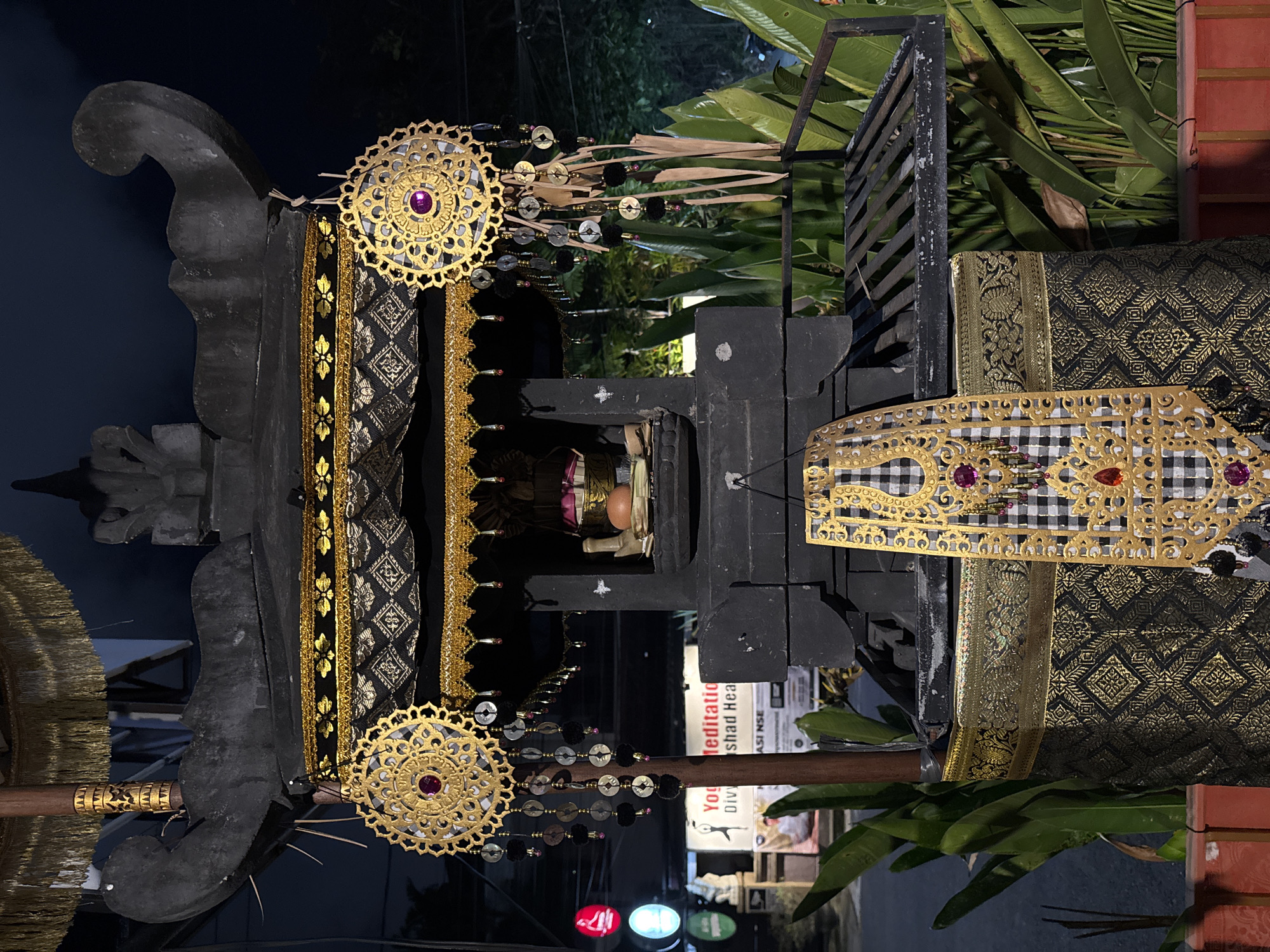 This media file depicts an elaborately decorated altar, likely for a religious or cultural ceremony, in Kuta, Indonesia. The altar is primarily black with ornate gold trim and dark, intricately patterned fabric. Large, circular gold ornaments with purple jewels are prominently featured on either side, adorned with hanging strings of beads and dark pom-poms. A checkered black and white fabric band, also edged with gold filigree and red jewels, is draped across the top. Green foliage is visible in the background, suggesting an outdoor or semi-outdoor setting. A wooden pole with gold accents extends from the left side of the altar. In the center of the altar, a small, enclosed space contains what appears to be a reddish-brown oval object, possibly an egg, and some other offerings. Beneath the altar, a sign is partially visible with text that appears to include "meditation", "You", and "Divi". The lighting suggests it is nighttime, with some colored lights (red and white) reflected below. The overall impression is one of a culturally significant and festive occasion.