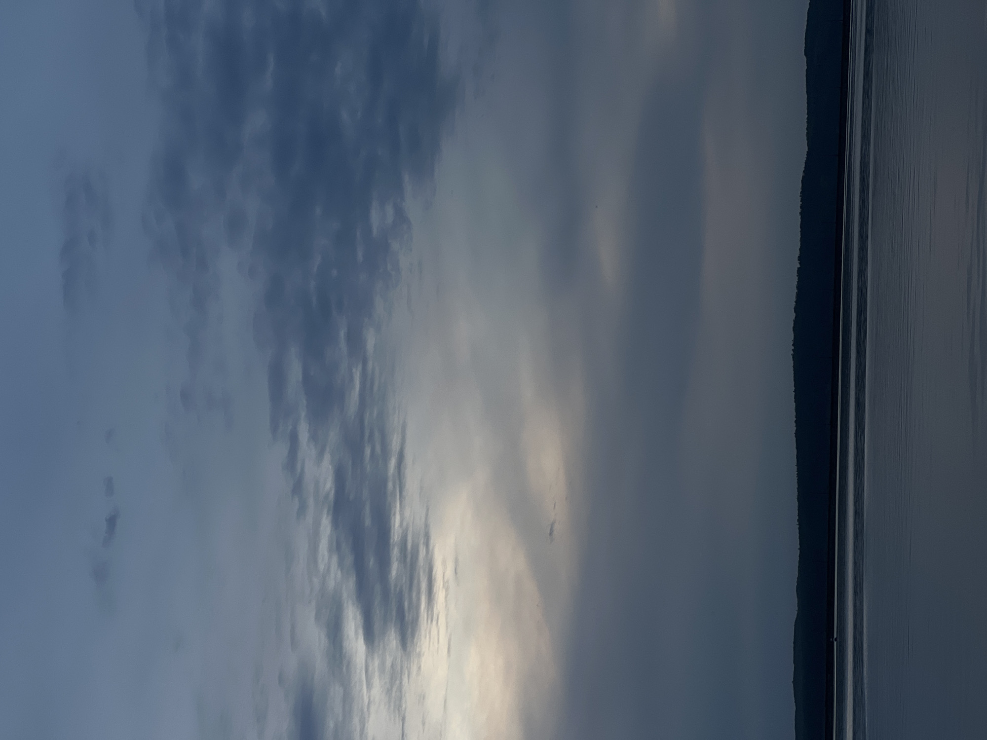 The image depicts a somber, overcast scene. The sky is dominated by dark, dense clouds with hints of a brighter area peeking through. The right side of the image shows a dark, treeline silhouette along the water's edge, suggesting a body of water. The location is likely Chaikovsky, Russia. The overall mood is gray and muted.