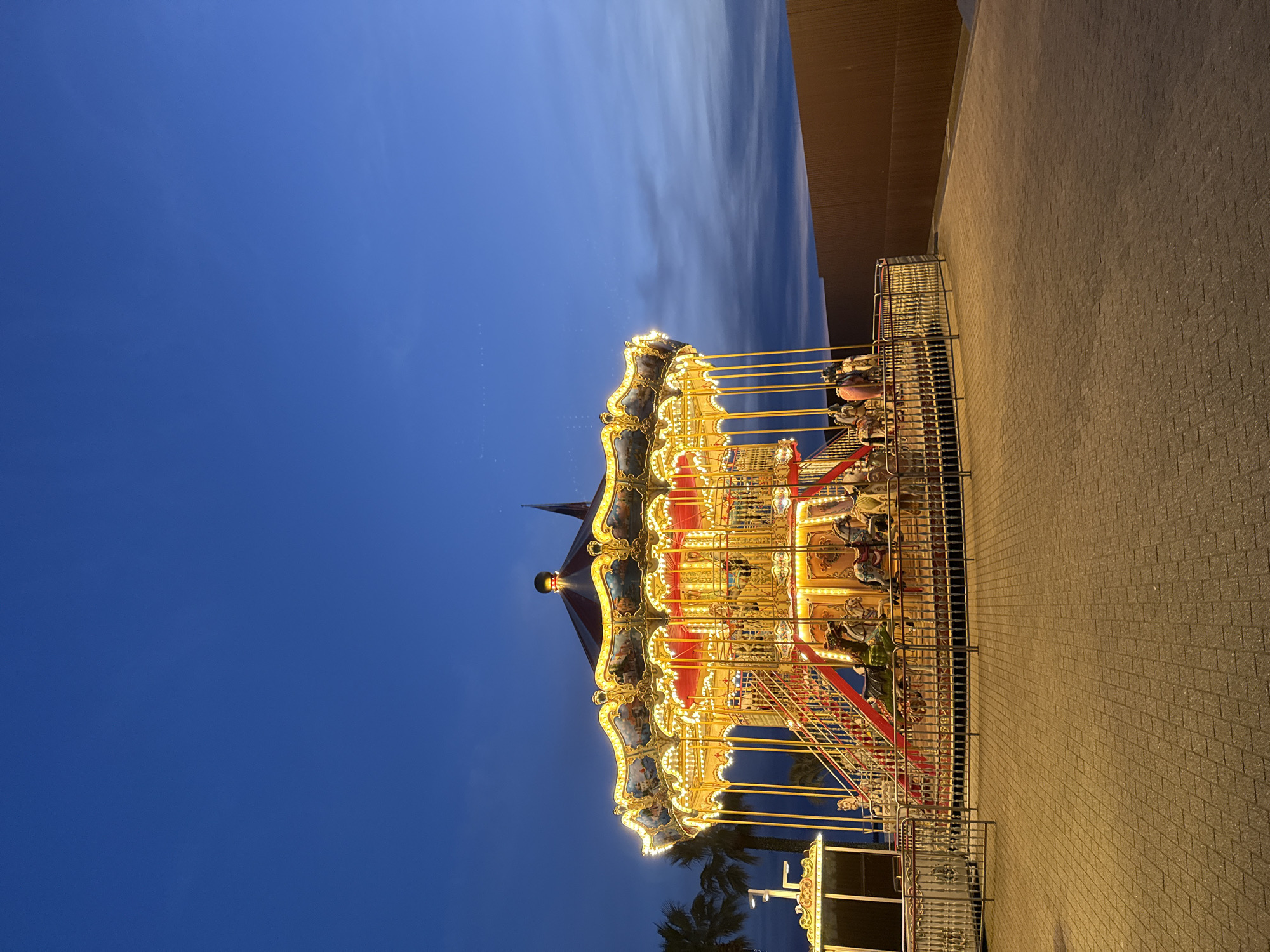 An illuminated carousel stands as the central subject in a vibrant nighttime scene. The carousel is richly decorated with numerous warm yellow lights outlining its multi-tiered structure and ornate golden details. Its roof is dark with red sections, and classic carousel figures, likely horses, are visible carrying indistinct riders.
The sky above and to the left of the carousel is a deep, clear twilight blue, hinting at either dusk or early night, with faint wispy clouds visible in the upper reaches. To the right, a large, nondescript building with a textured brown-beige wall and a darker brown roof provides a backdrop. The ground in the foreground and extending to the right is a wide area of light-colored, possibly brick or cobblestone paving. Metal railings enclose the carousel's base. Silhouettes of palm trees can be seen near the bottom of the carousel, suggesting a warm climate. The scene depicts an active entertainment setting, likely an amusement park or public square. No discernible text is visible in the image.