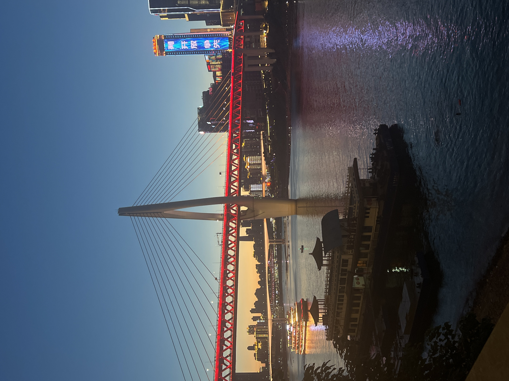 An evening shot of the Yangtze River in Yuzhong District, Chongqing, China. A large cable-stayed bridge with red lighting spans the river. Two multi-story passenger boats are docked in the foreground. The cityscape, illuminated by lights, is visible in the background. The sky is a deep blue, transitioning to a warmer orange hue near the horizon. The water reflects the colorful lights of the bridge and buildings.