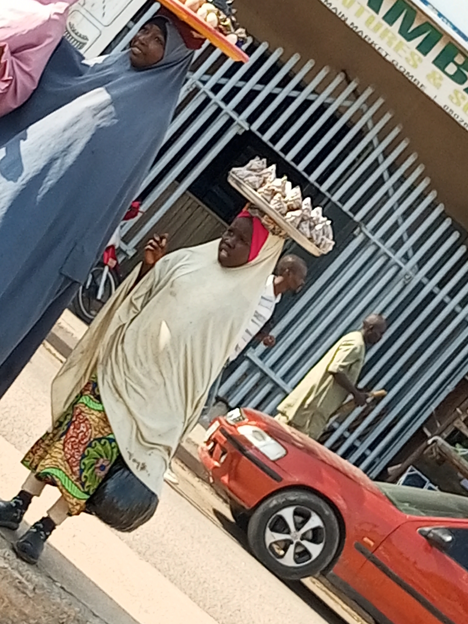 In Arawa, Nigeria, two women and two men are depicted outdoors. A woman on the left is partially visible, wearing a pink garment and a long blue abaya. To her right, another woman is prominently featured, balancing a tray of goods on her head. She is wearing a light-colored robe and a patterned skirt. A third person, a man, is seen in the background, wearing a green shirt. Another man is partially visible in the mid-ground, wearing a white shirt. A red car is parked in the foreground. The scene appears to be set along a street, with a building and metal fencing in the background.