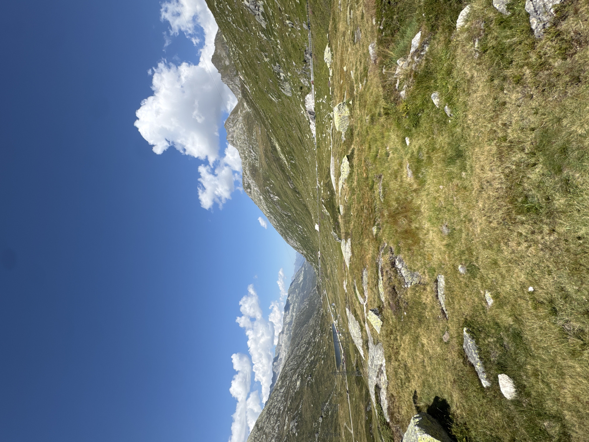The image depicts a panoramic view of a mountainous landscape, likely in the vicinity of Airolo, Switzerland, characterized by lush green slopes, rocky terrain, a winding road, and a small body of water. The sky is a vibrant blue, dotted with scattered white clouds. The foreground is dominated by a grassy meadow with numerous large, light-colored rocks. In the mid-ground, a road snakes through the valley, bordered by a small, dark blue lake on the left. The background features imposing, rugged mountains, their slopes covered in green vegetation and interspersed with rocky outcrops. The overall impression is one of natural grandeur and a serene, alpine environment. There are no visible people or specific man-made structures that would pinpoint a precise location within Airolo, beyond the general mountainous setting.