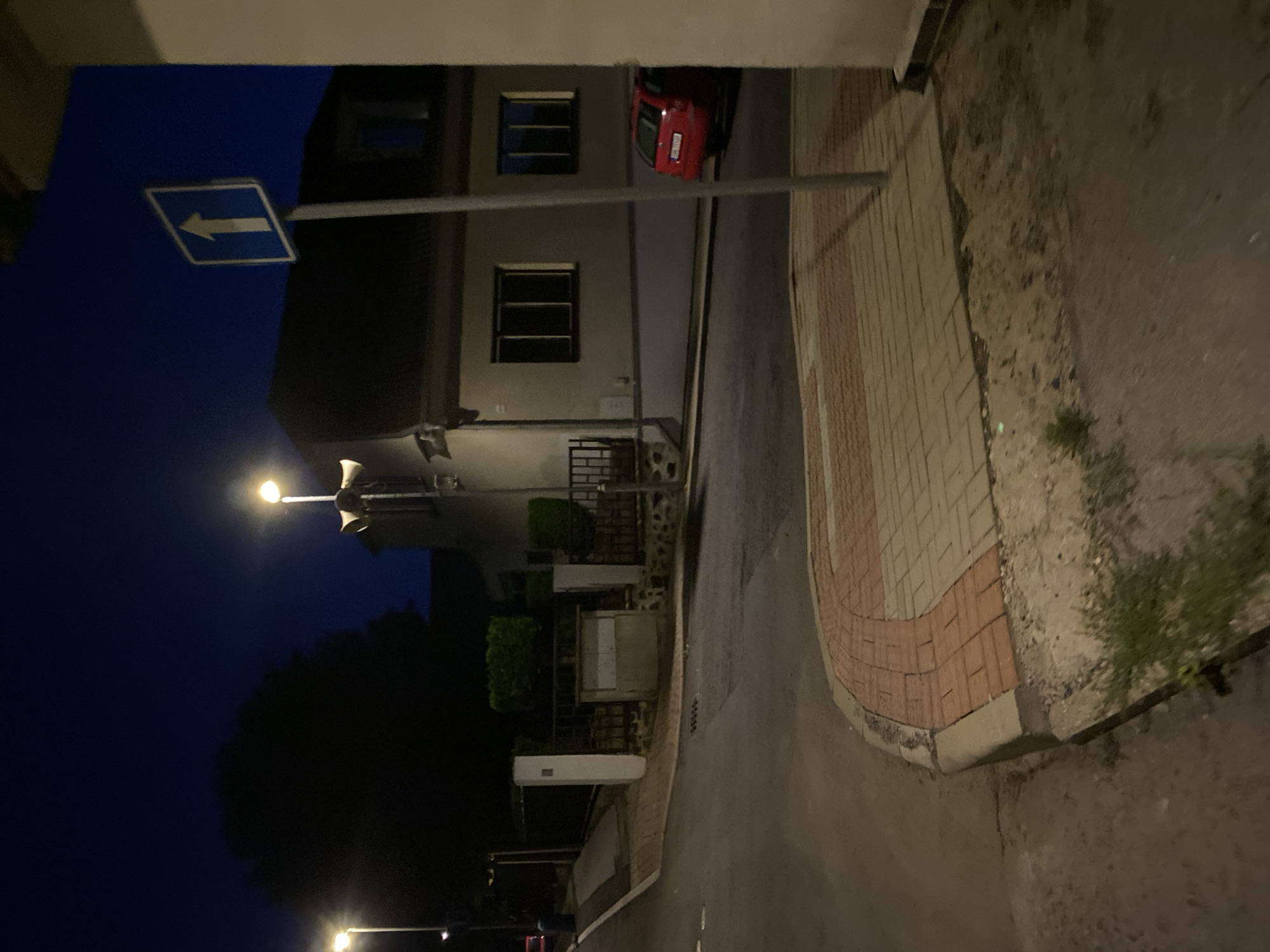 The image depicts a street scene at night in Bílina, Czechia. A street lamp illuminates a pathway and a section of a building. A white directional sign with an upward-pointing arrow is visible. Adjacent to the sign, a building with dark windows is present. A red car is parked on the street. The street is paved, and a sidewalk area with interlocking brick paving is also visible. In the foreground, there is unkempt vegetation and a concrete curb. The sky is dark blue, indicating it is nighttime.