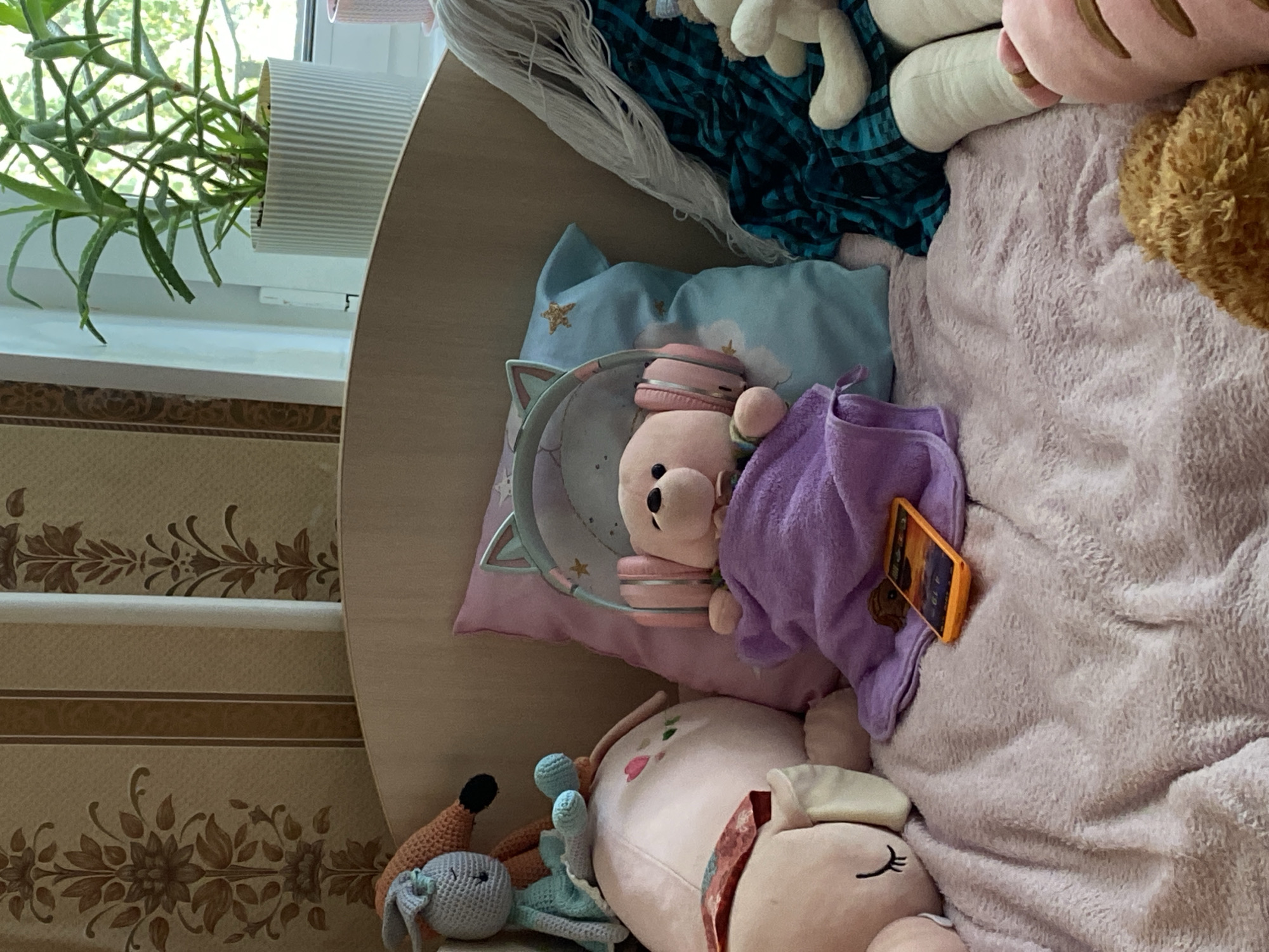 The image depicts a cozy bed with several stuffed animals, suggesting a child's bedroom. The stuffed animals include a blue crocheted bunny, a pink teddy bear wearing headphones, a large pink stuffed animal, and other unidentified plush toys. A smartphone is placed on a purple blanket on the bed. The background shows floral wallpaper and a window with a potted plant, indicating an indoor setting. The presence of a potted aloe vera plant on the windowsill suggests a location with sufficient natural light. The overall impression is one of comfort and domesticity.
It is not possible to determine the specific location within Lomonosov, Russia, based solely on the visual information provided in the image. The image shows a bedroom interior with common household items and decor. There are no landmarks, street signs, or other contextual clues that would allow for precise geographical identification within Lomonosov or any other city.