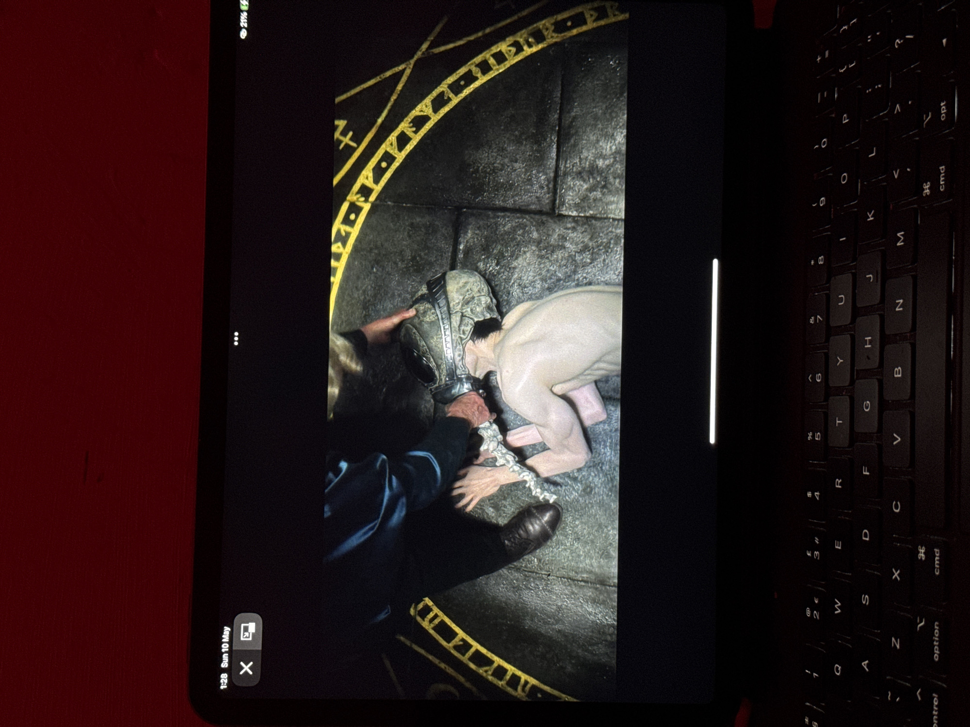 A photograph of a scene being displayed on a tablet, captured on what appears to be a red surface, with a black keyboard to the right. The tablet's display shows a person putting a helmet or mask on another individual, who is kneeling on a stone surface inscribed with a yellow geometric pattern. The person applying the mask is wearing a dark blue or black jacket and brown shoes. There is also a chain involved. The date "Sun 10 May" and a battery indicator showing "21%" are visible on the tablet screen. The city of Sofia, Bulgaria, is not discernable from this image.