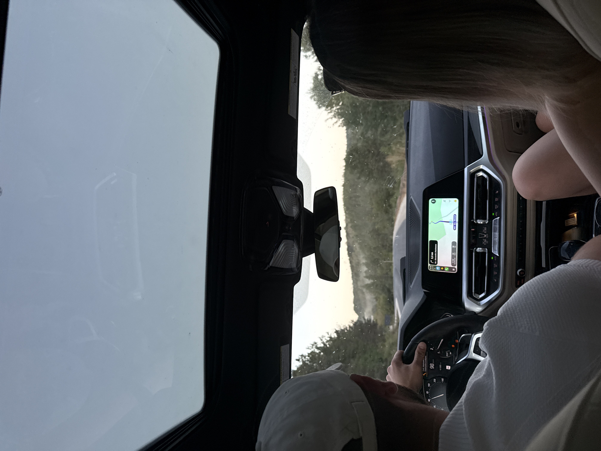 The image shows the interior of a car, looking forward through the windshield and sunroof. Two people are visible, a male driver in a white cap and a female passenger in the front seat. The driver's hands are on the steering wheel, and the car's dashboard and infotainment screen are visible. The infotainment screen displays a navigation map with a route leading to Sărata-Mereșeni, Moldova. The car is on a road surrounded by trees and hills, suggesting a rural or mountainous landscape. The sky above is partly cloudy and the lighting suggests it is either early morning or late afternoon. There is no indication of the exact location within Sărata-Mereșeni itself, beyond it being the destination of the car's navigation.