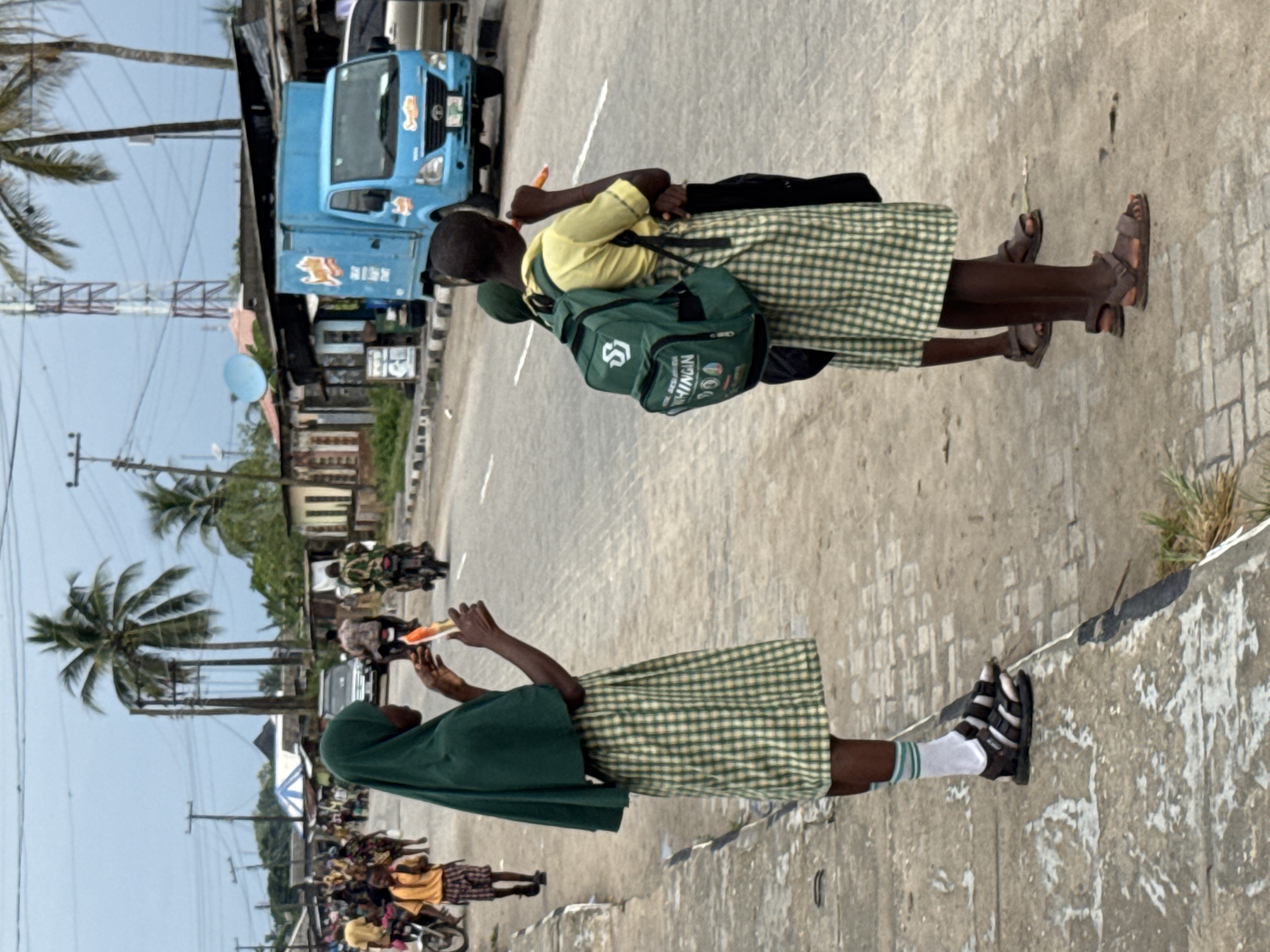 The image depicts school children in Badagry, Nigeria. The location is on an urban street with vehicles, including a blue truck, and motorcycles. Some children wear school uniforms consisting of green and yellow plaid skirts, with some wearing yellow shirts and green headscarves. Some of the students are on the road’s edge, while others are walking on the sidewalk. The students wear sandals. The environment features palm trees and utility poles.
