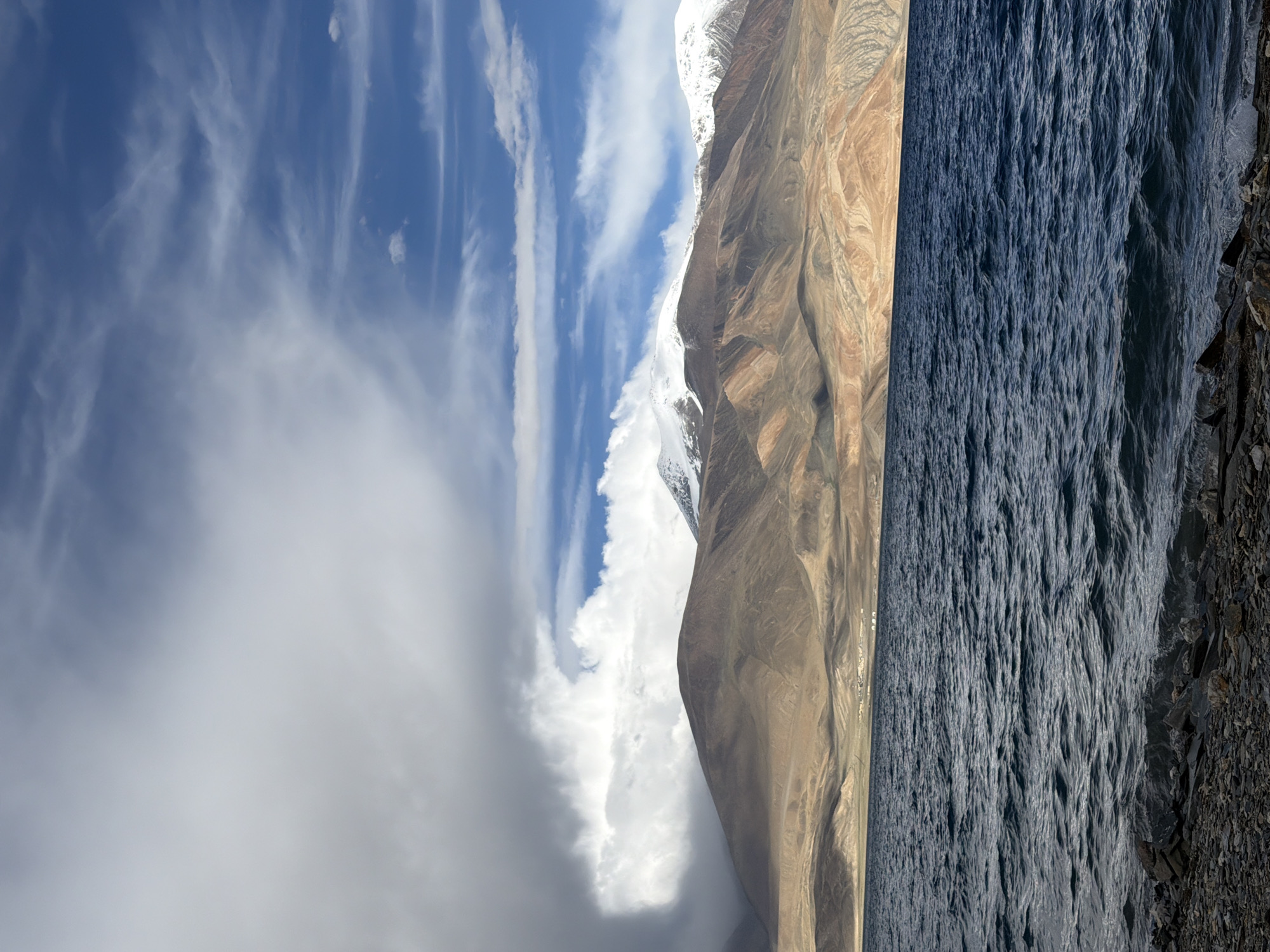 A wide-angle, full shot captures a serene body of dark blue water in the foreground, gently lapping at a rocky shoreline. Behind the water, a vast, arid mountain range stretches across the horizon, its slopes a mix of earthy browns and ochres. Snow-capped peaks are visible in the distance, adding a stark contrast to the dry landscape. The sky above is a dramatic canvas of blues and whites, with large, textured clouds dominating the upper half of the frame. Some clouds are dark and brooding, while others are wispy and translucent, catching the sunlight. The scene suggests a remote, natural landscape, possibly in a high-altitude region of India. No people or man-made structures are discernible in the immediate vicinity.