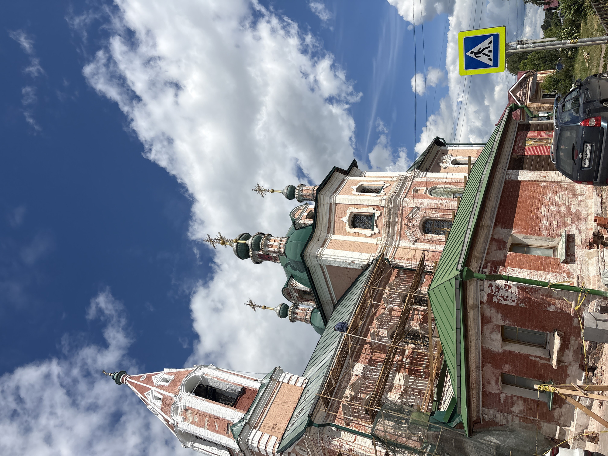 This image depicts an exterior view of a historic Russian Orthodox church under renovation. The church features traditional architecture with red brick walls, white decorative elements, and multiple green-domed cupolas topped with golden crosses. Scaffolding is visible along one side of the building, indicating ongoing repair work. The sky is a vibrant blue, with fluffy white clouds scattered across it, suggesting a bright, possibly daytime scene. A yellow pedestrian crossing sign is prominently displayed on a pole on the right side of the image. In the background, trees and other buildings are visible. A dark-colored car is parked near the church. The overall scene conveys a sense of historical preservation and community life. No people are clearly visible in the image.